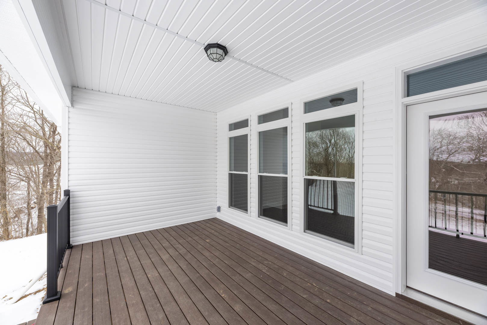 White exterior home with large windows, wood deck flooring, and metal siding; ceiling light fixture visible inside; tree branches seen through window.