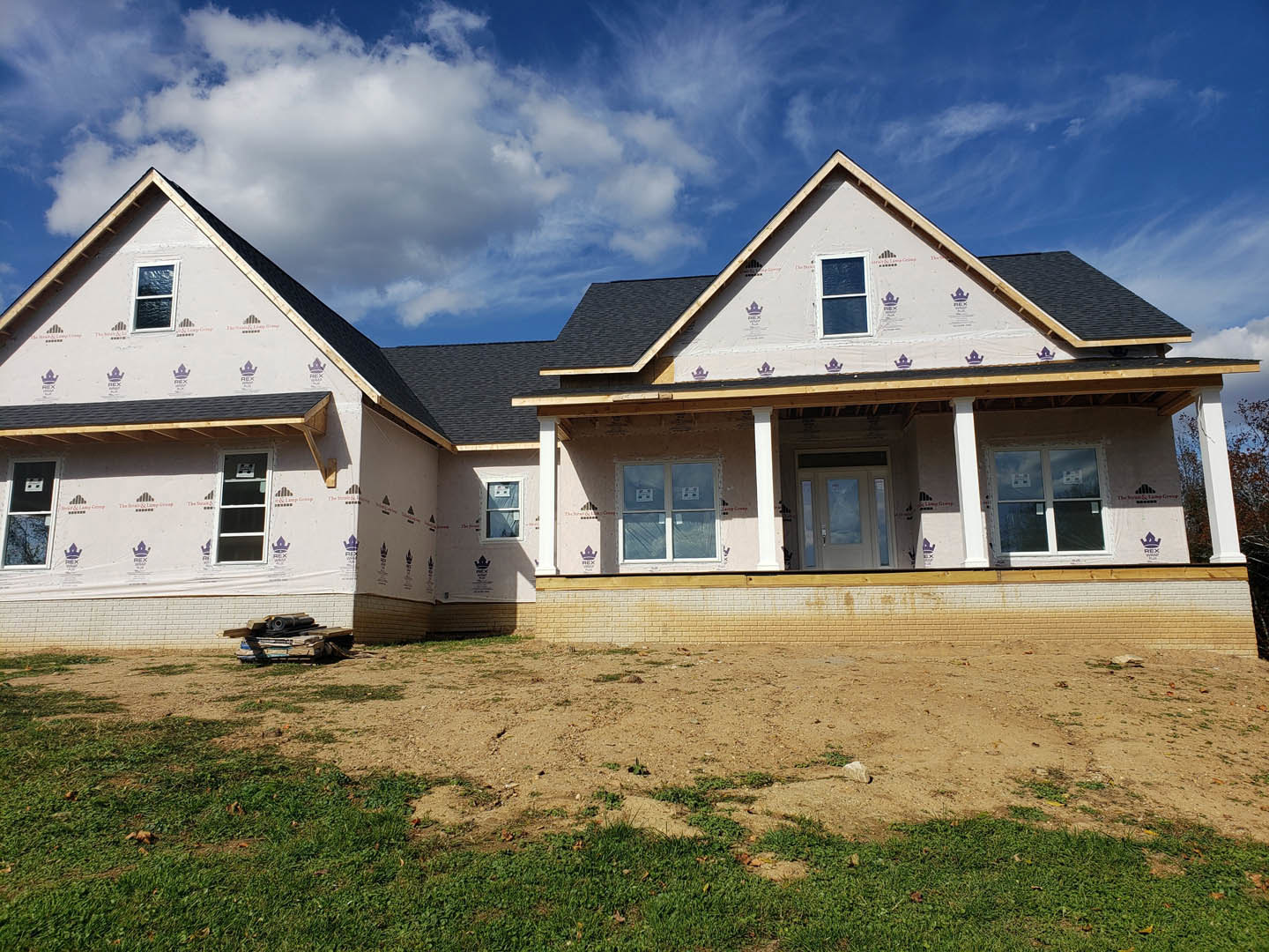 House under construction with exposed framing, dirt field in foreground, white-framed window and door, American Gothic House visible in background under blue sky