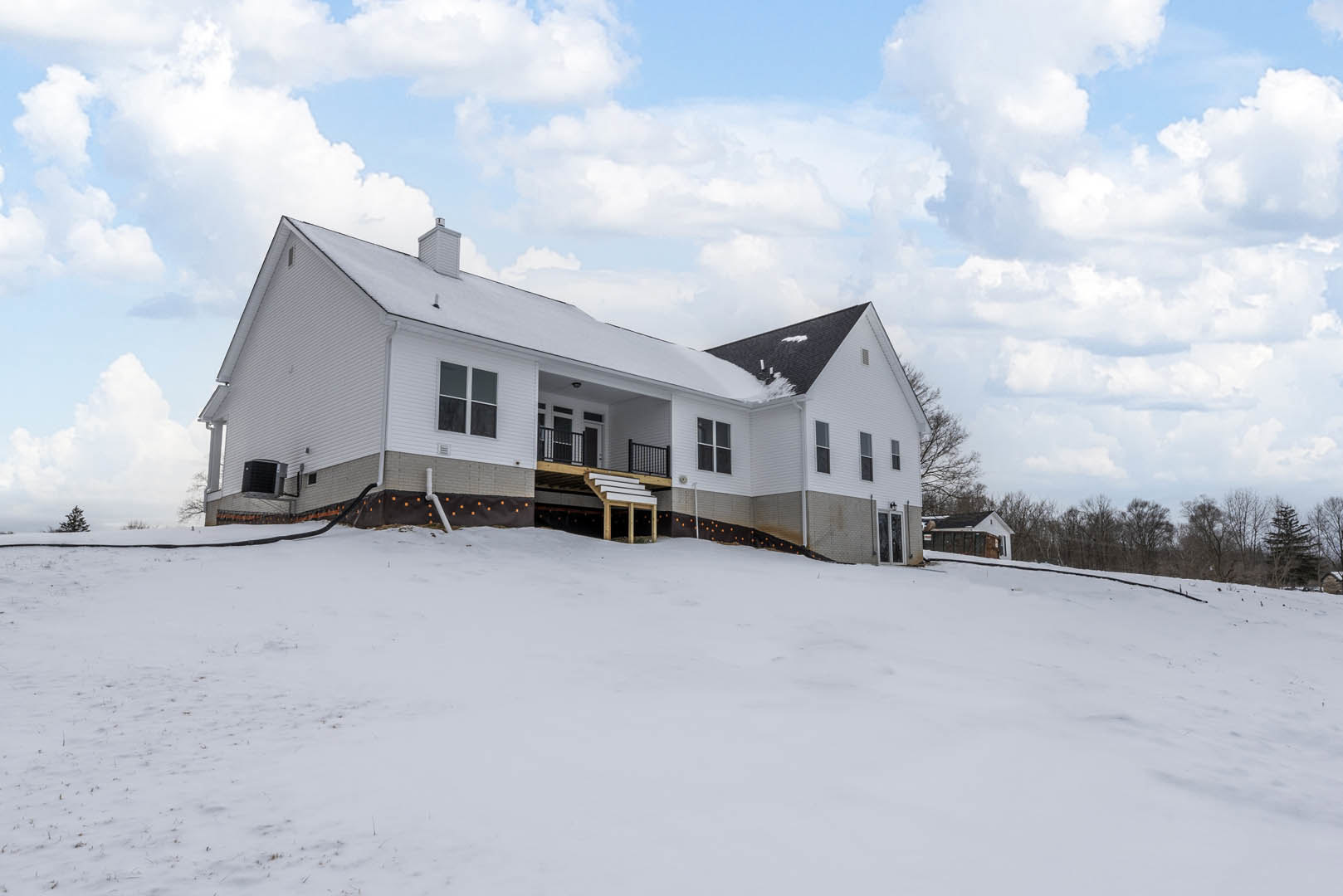 White house with snow on the roof, covered porch, wooden deck with stairs, white chimney, surrounded by snow-covered hill and bare trees under cloudy winter sky