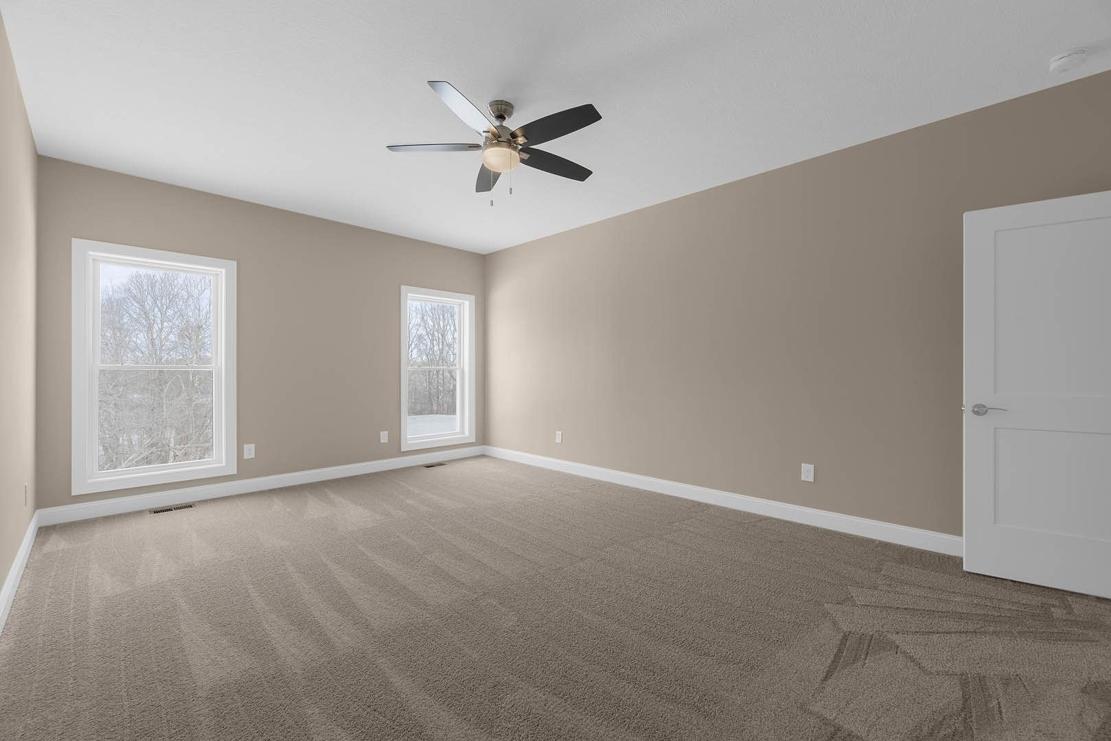 Neutral-toned room with carpet flooring, ceiling fan with light fixture, large windows showing leafy trees, and white door with silver handle
