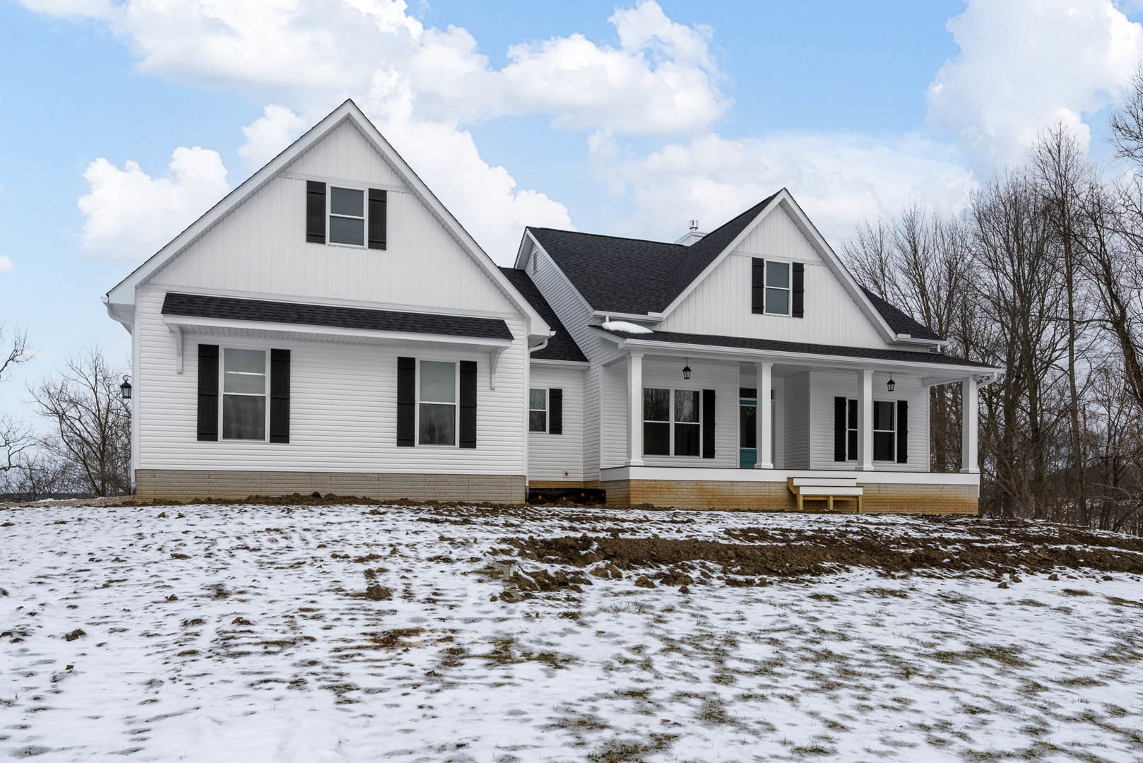 White house with black shutters and white-framed windows, snow covering the ground, bare trees nearby, cloudy sky overhead