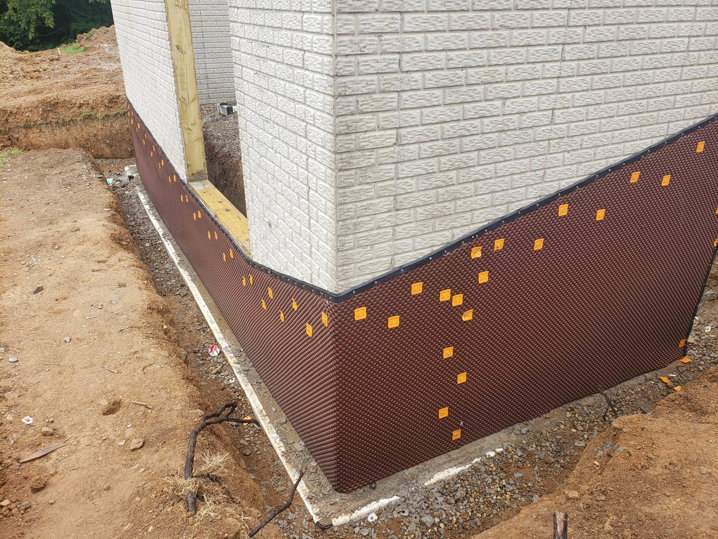 Red and brown brick wall with a hole in the corner, brick base, green tree and bushes nearby, tree branch on the ground, dirt and metal fence in the background