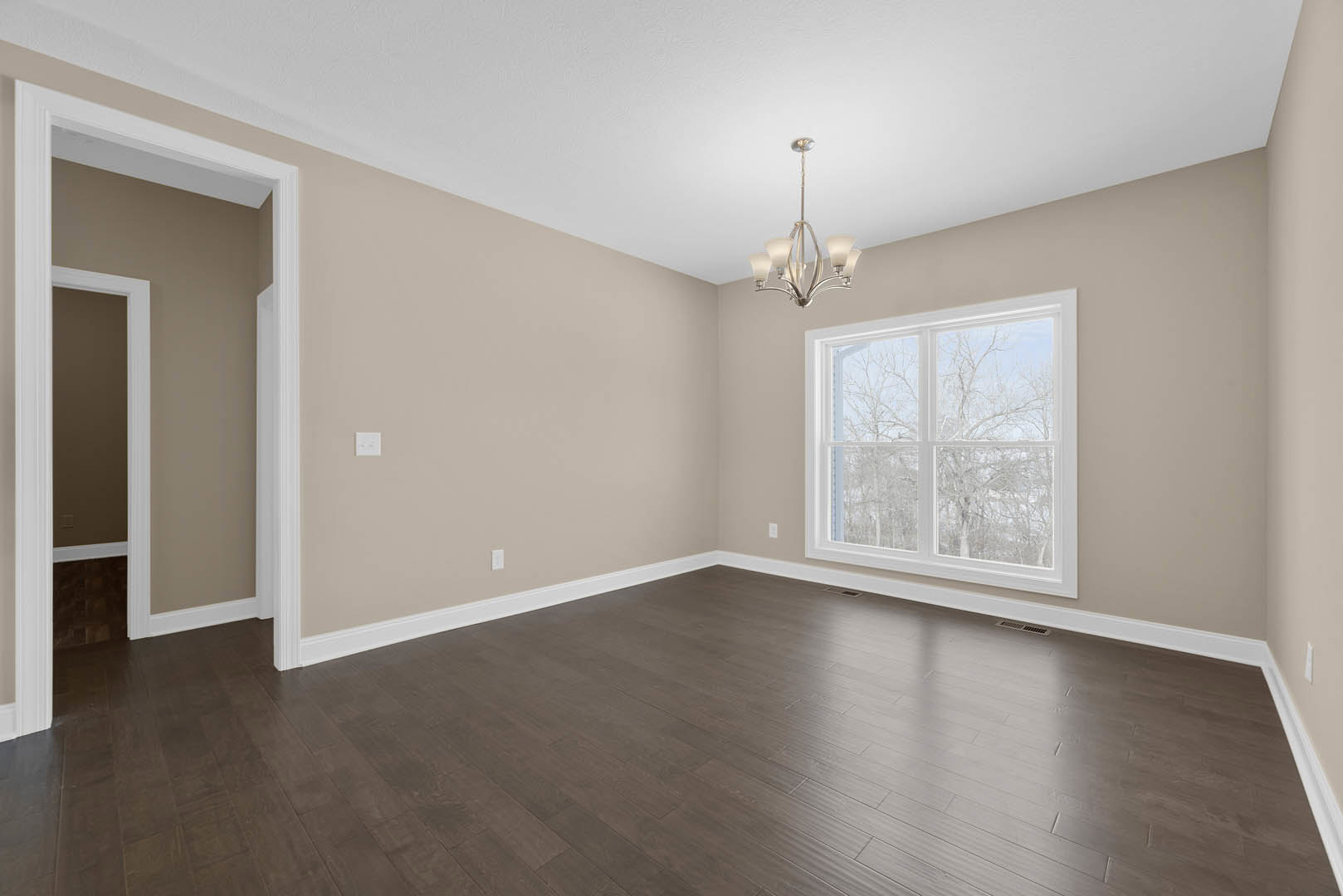 Sunlit room with large window overlooking trees, hardwood flooring, white framed doorway, white plaster ceiling with modern chandelier