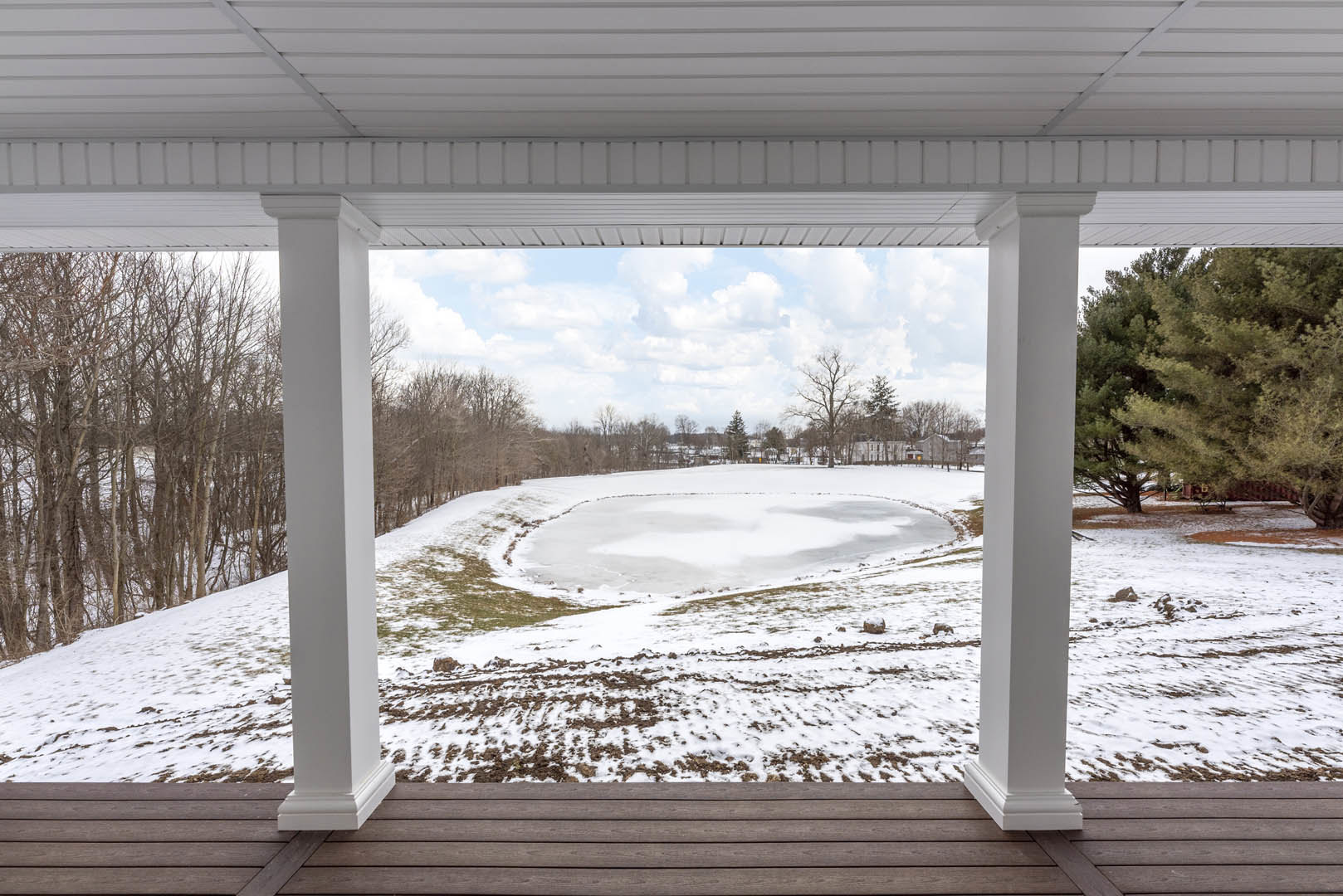 Wooden covered porch with white ceiling and pillars, snow-covered deck, frozen pond and leafless trees in winter landscape