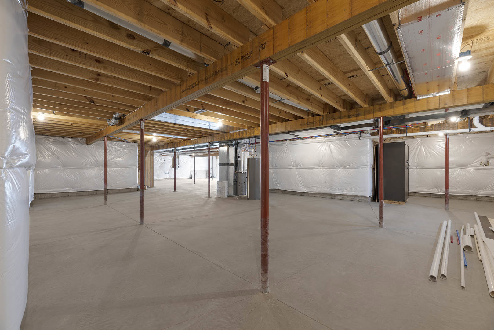 Exposed wooden ceiling beams and a steel support pole in a basement room with white wall insulation and unfinished concrete floor
