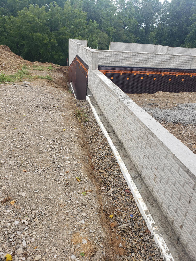 White exterior wall with white brick and brown-and-white brick sections, adjacent to dirt and gravel ground, curb, and construction materials.