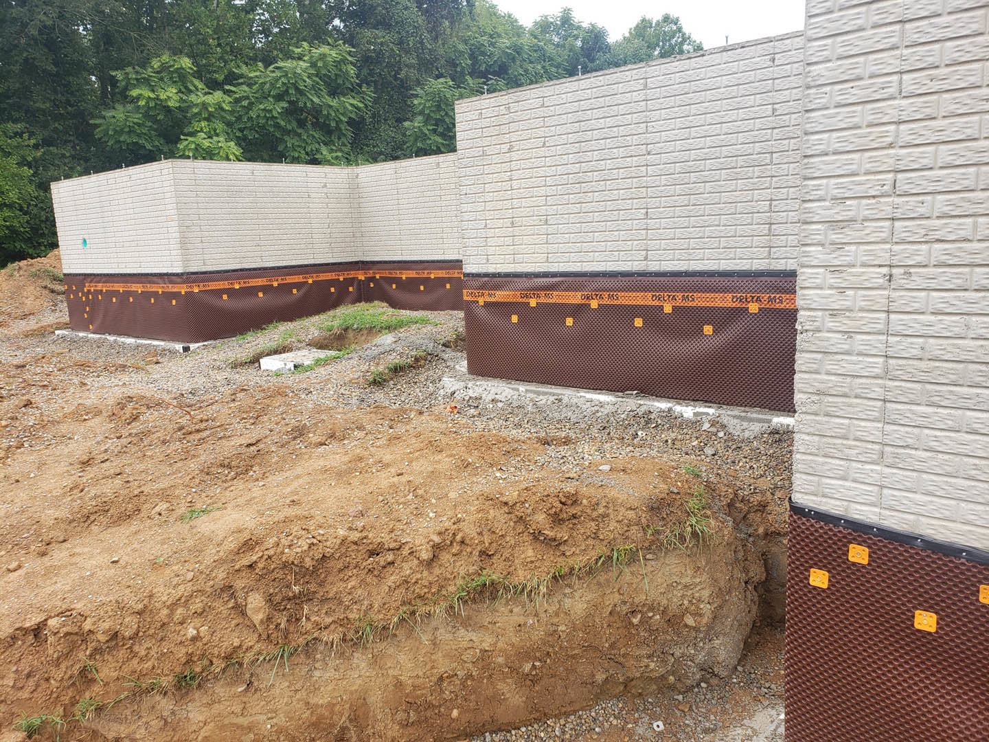 Brown brick exterior wall with a rectangular hole in the ground, surrounded by dirt, orange and black construction materials, and patches of grass.