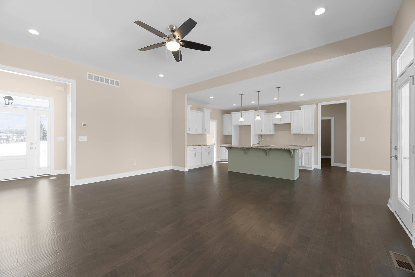 Spacious open floor plan featuring dark wood laminate flooring, central kitchen island with light countertop and shelf, ceiling fan with integrated light, white plaster walls, and