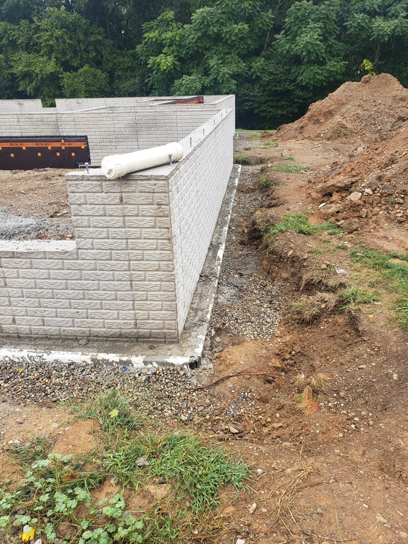 Construction site with exposed soil piles, white plumbing pipe along concrete foundation, black and orange protective fabric, scattered rocks and grass, partially built structure
