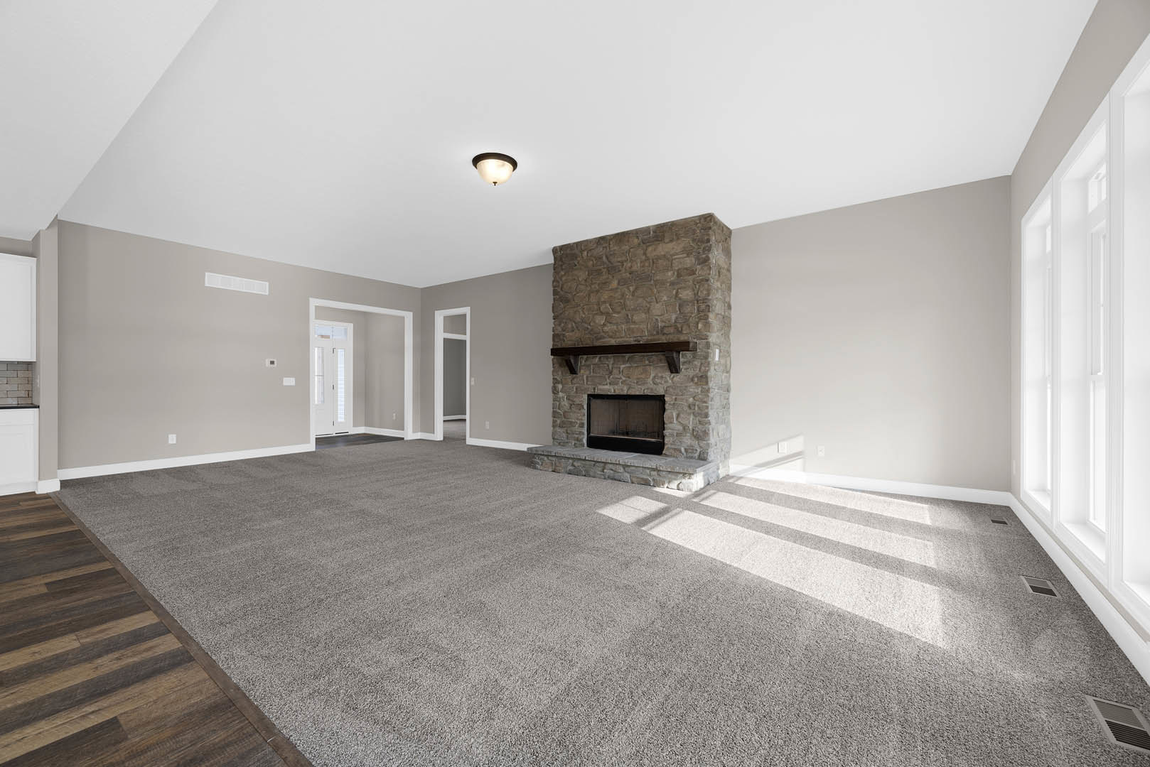 Living room featuring a stone fireplace with a wood mantel, carpeted floor transitioning to wood flooring, white-shaded ceiling light, and plaster walls.
