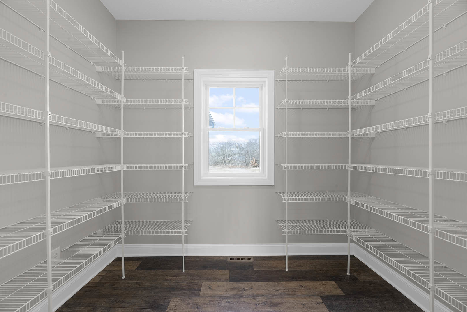 White wire shelving unit along a plaster wall, wood floor with vent, large window overlooking snowy landscape and trees under blue sky