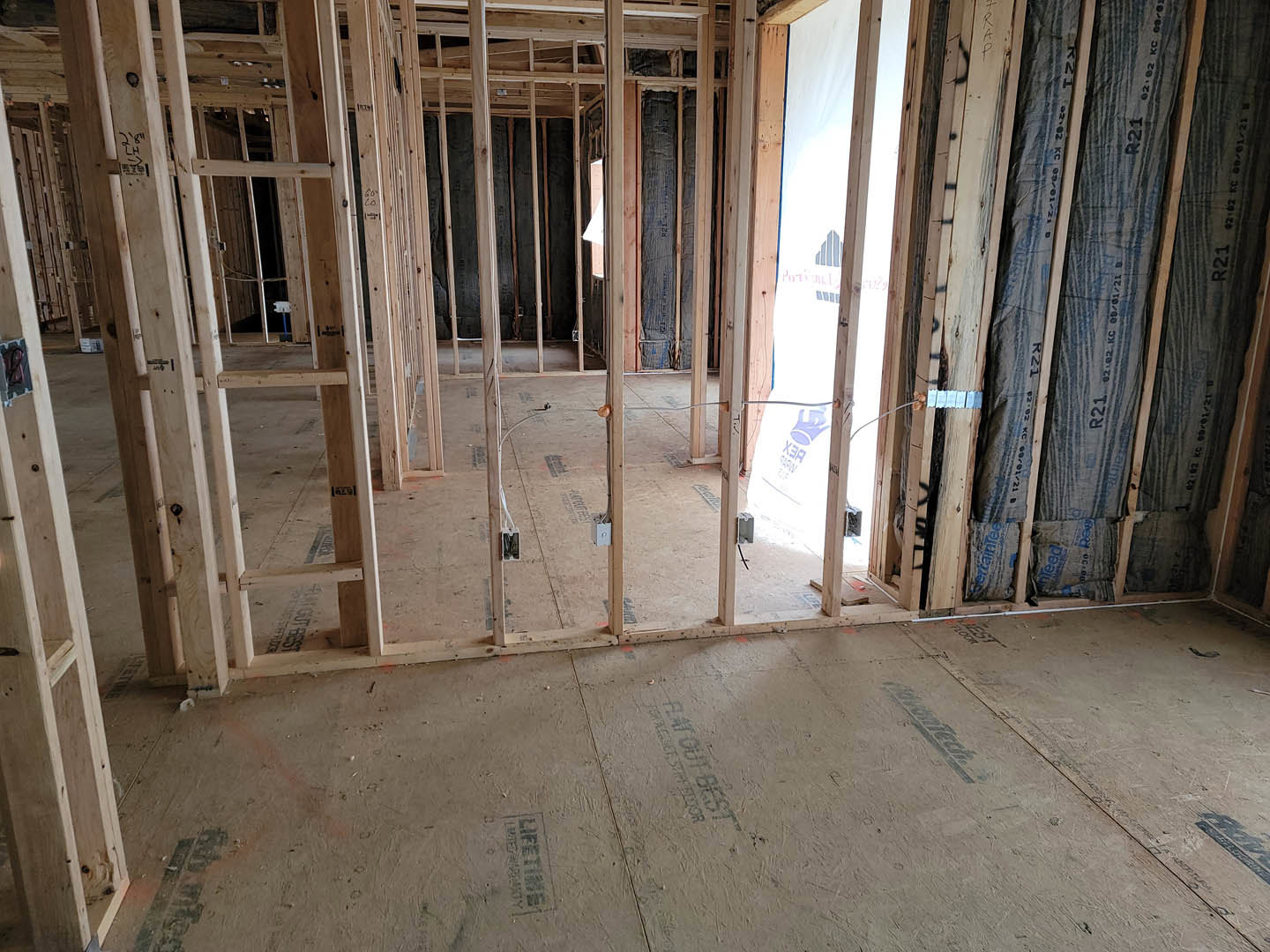 Unfinished room with exposed wood framing, subfloor panels marked with text, window frame displaying a logo, construction bag, wood beams partially wrapped in black material, and