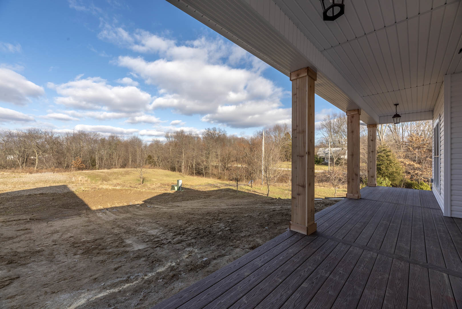 Wooden porch with square pillars overlooking a dirt field, scattered trees in the background, cloudy sky above, white utility box visible in the field