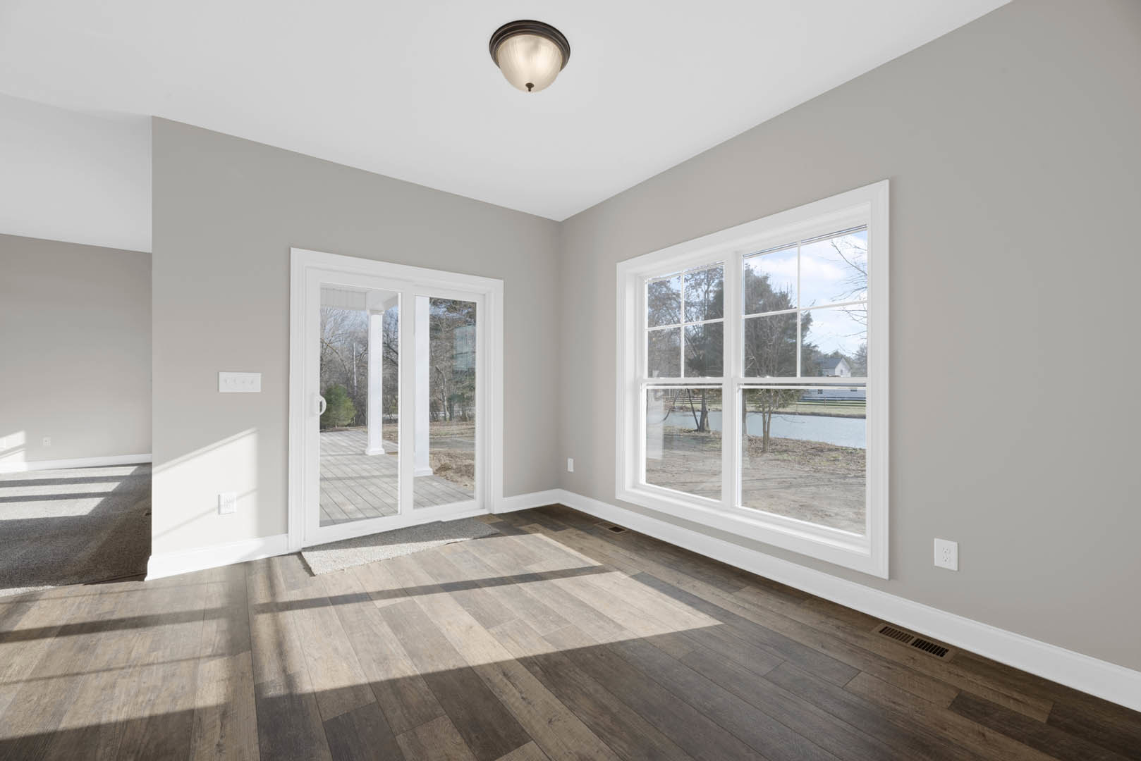 Open room with wide plank wood flooring, large windows overlooking river and trees, white door with glass panels, neutral walls with crown molding, modern light fixture, area rug