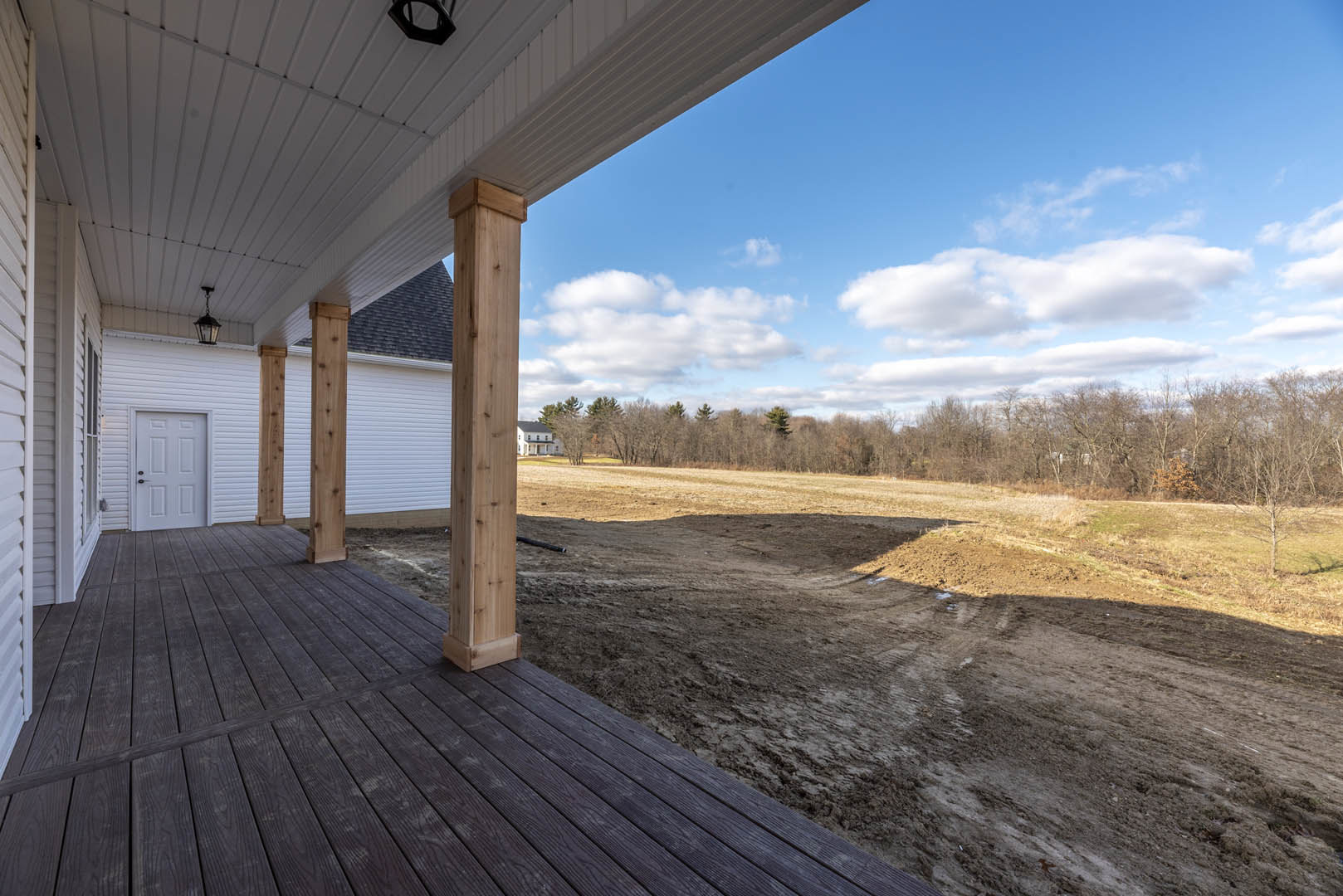 Modern home with white door and black handles, wooden porch with pillars, wood panel featuring holes, expansive dirt field in foreground, cloudy sky, sparse trees, and grassy