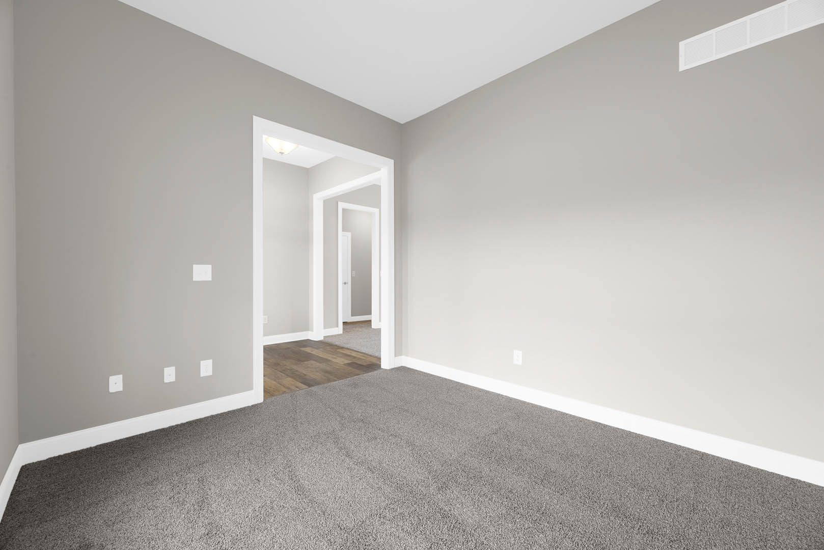 Grey carpeted floor and white walls with a white door open in a white frame, brown laminate flooring visible beyond doorway.