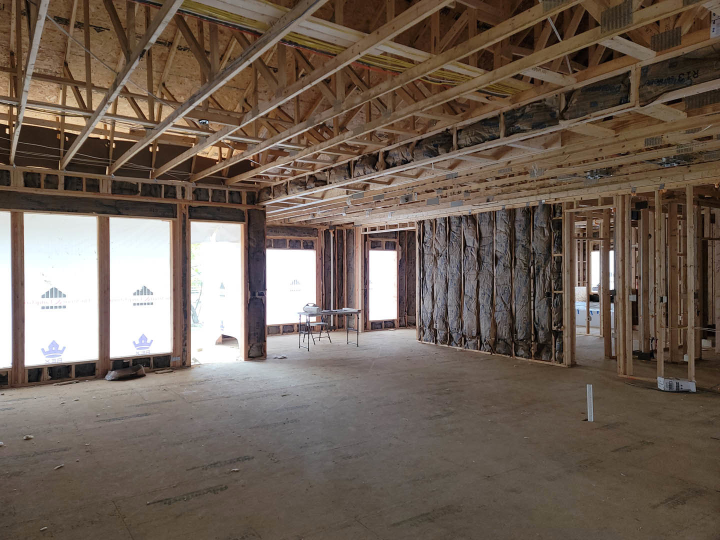 Open-concept room with exposed wooden ceiling beams, large windows with white coverings, light-colored walls, and a mix of chairs and tables; bags stacked against one wall.