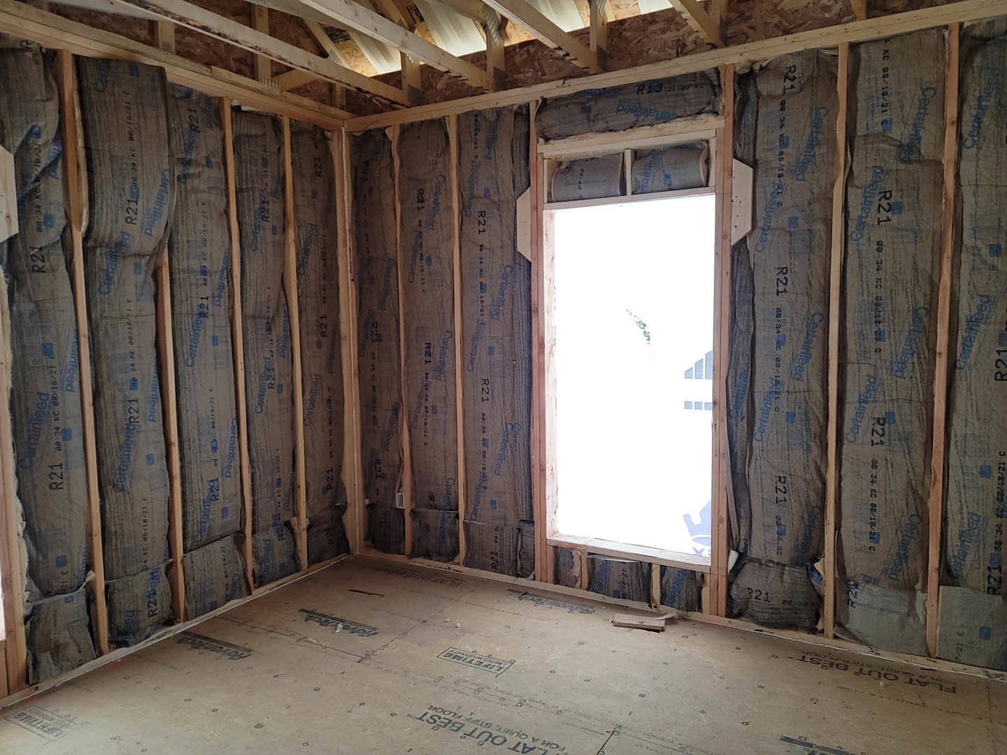Living room with exposed wood ceiling beams, large window showing blue sky, light-colored walls, and floor featuring black lettering.