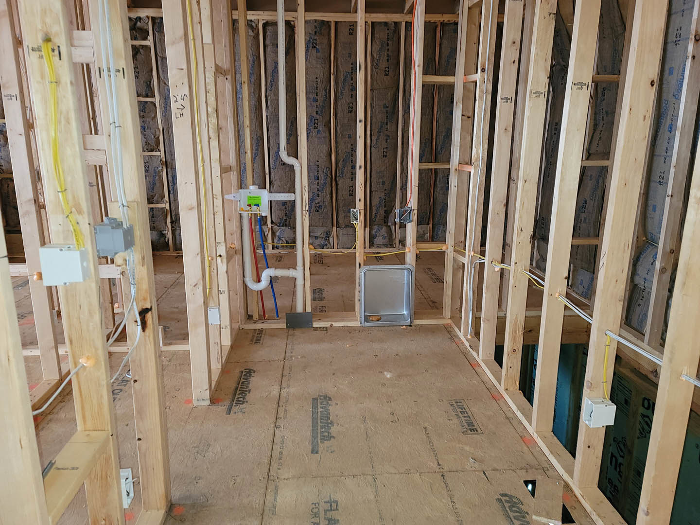 Exposed wood framing and beams with visible plumbing pipes, electrical boxes, and construction materials inside a residential home under renovation