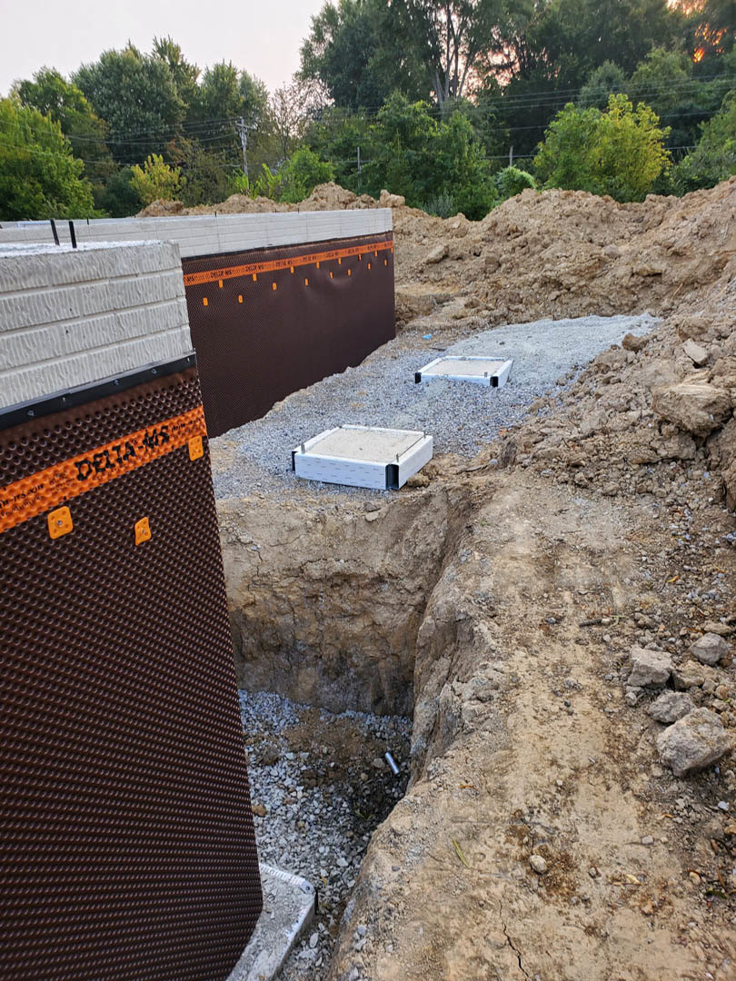 Construction site with exposed dirt and rocks, concrete blocks, partially built wall, and surrounding trees under open sky