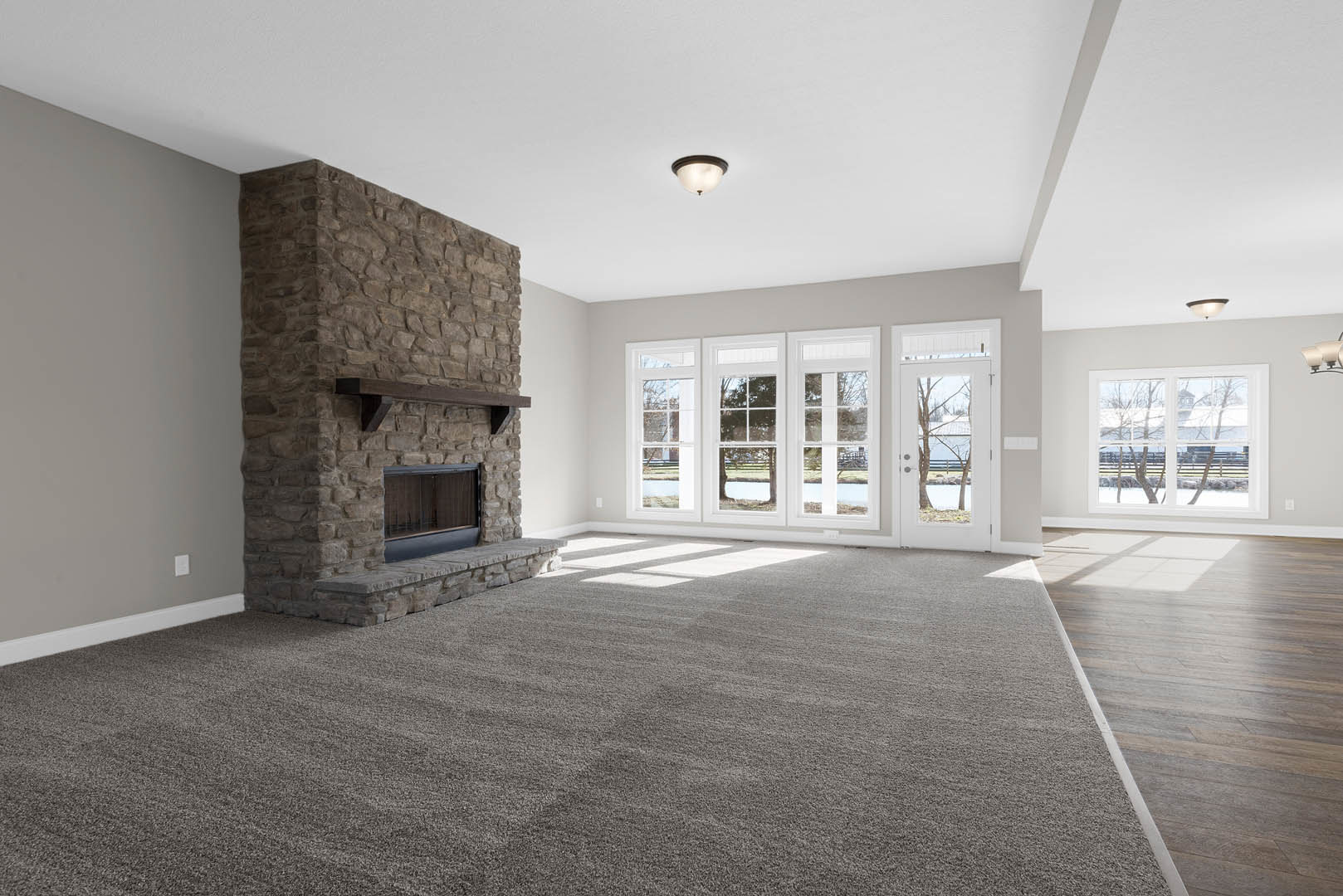 Living room with beige carpet, stone fireplace topped by a wood mantel, white-shaded ceiling light, plaster walls, large window overlooking farmland, partial view of metal gate