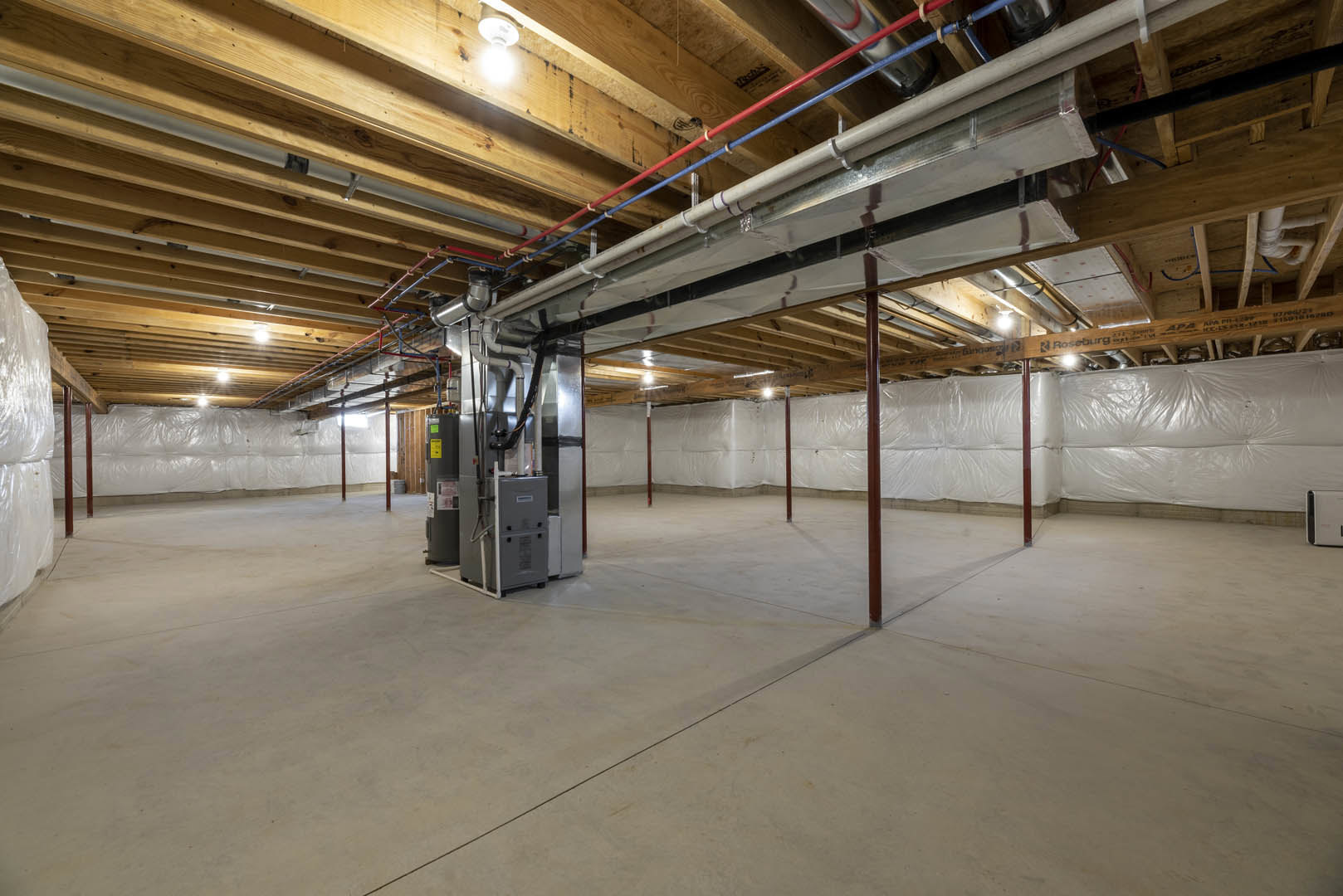 Basement room with exposed steel pipes along ceiling, concrete floor with central seam, single light bulb fixture, stack of white bags, and grey electrical box mounted on wall