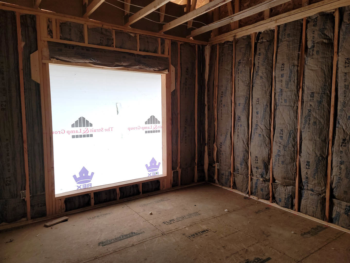 Room under construction with exposed wall insulation, window framed in white, unfinished wood surfaces, and subfloor marked with black text