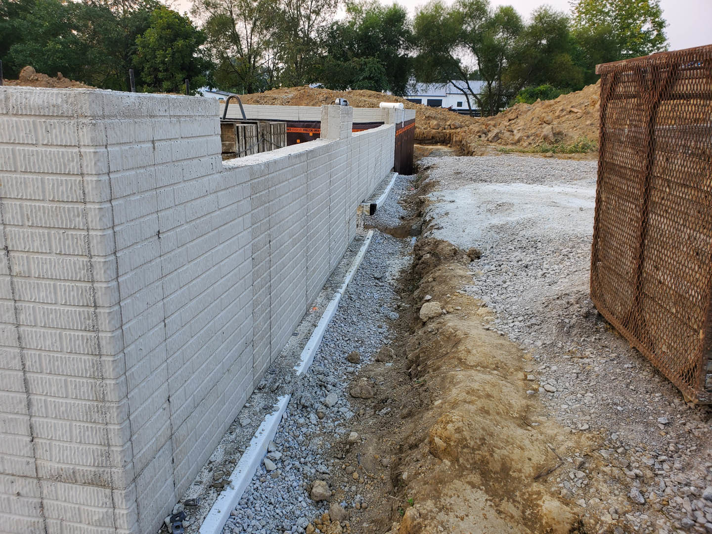 Red brick exterior wall with a large hole in the ground, surrounded by dirt, rocks, and a wooden fence; trees and sky visible in the background