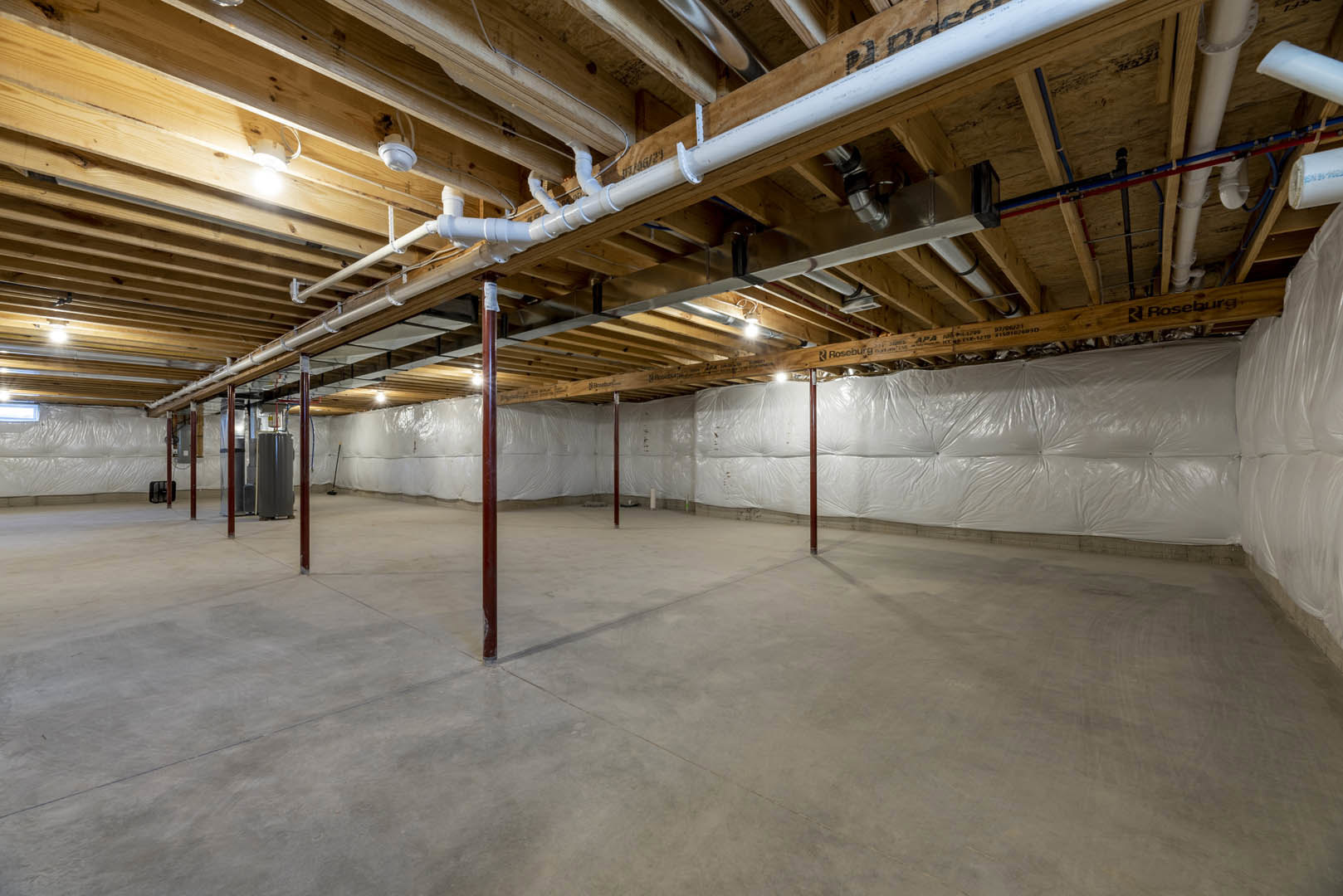 Concrete basement floor with white vapor barrier, exposed steel support pole, ceiling lined with blue, red, and white plumbing pipes.