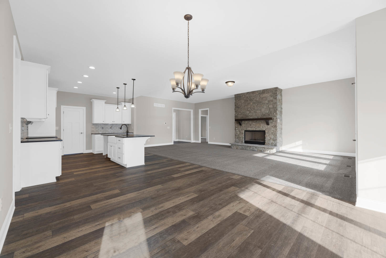 Spacious open floor plan living room featuring hardwood floors, stone fireplace with glass door, white kitchen island with drawers, white door with silver handle, and three-light