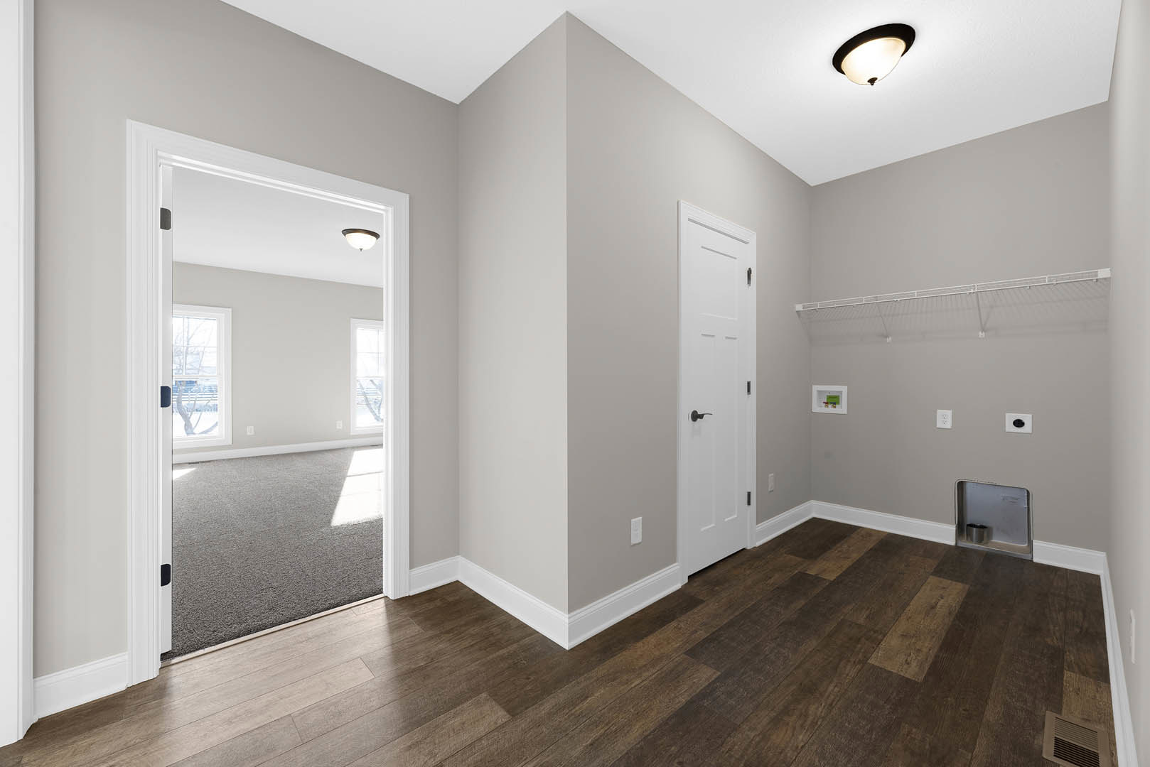 Entryway with white door and black handle, wood flooring and trim, white walls, coat rack, metal container recessed in wall, close-up light fixture, framed green and blue sign.