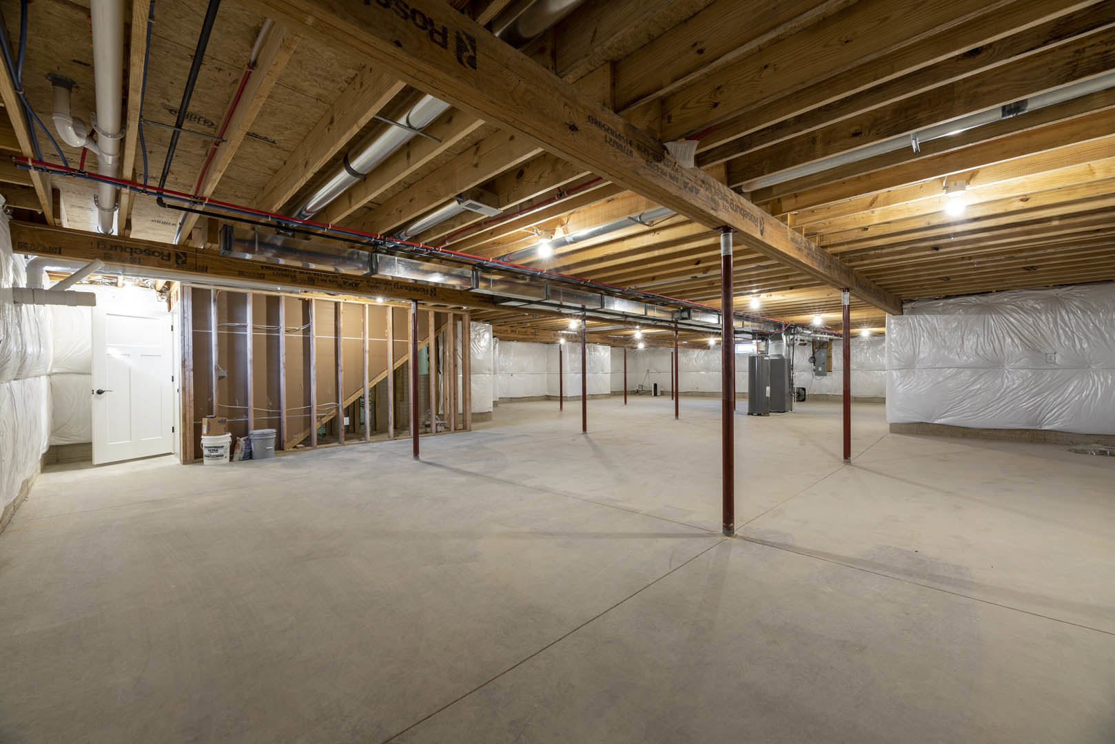 Exposed wooden ceiling beams and metal pipes above a concrete floor, red support poles, white plastic bag and bucket with blue label on a wooden surface in an unfinished basement