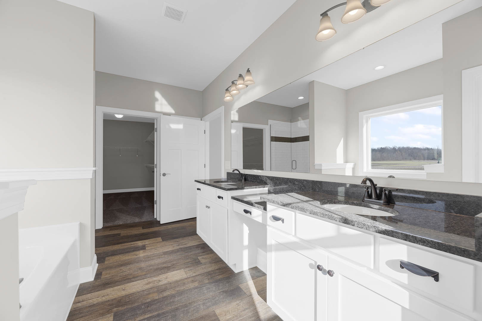 Bathroom featuring white cabinets, black countertops, under-mount sink, silver faucet, white door with silver handle, row of wall-mounted lights, window overlooking field and