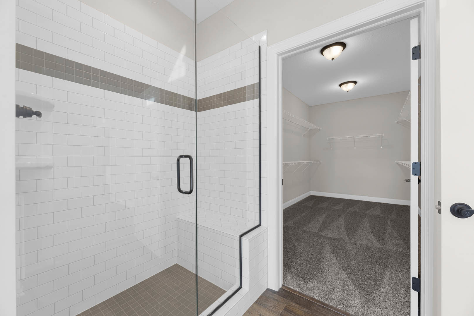 Bathroom with glass shower door, white tile walls, black shower handle, carpeted floor, ceiling light fixture, adjacent closet with shelves, window with white trim