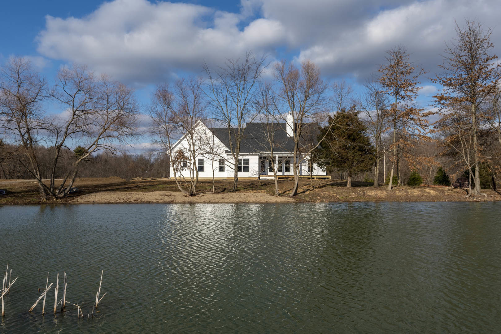White two-story house with large windows and gabled roof, surrounded by green trees, situated beside a calm lake with cloudy sky reflected in the water.