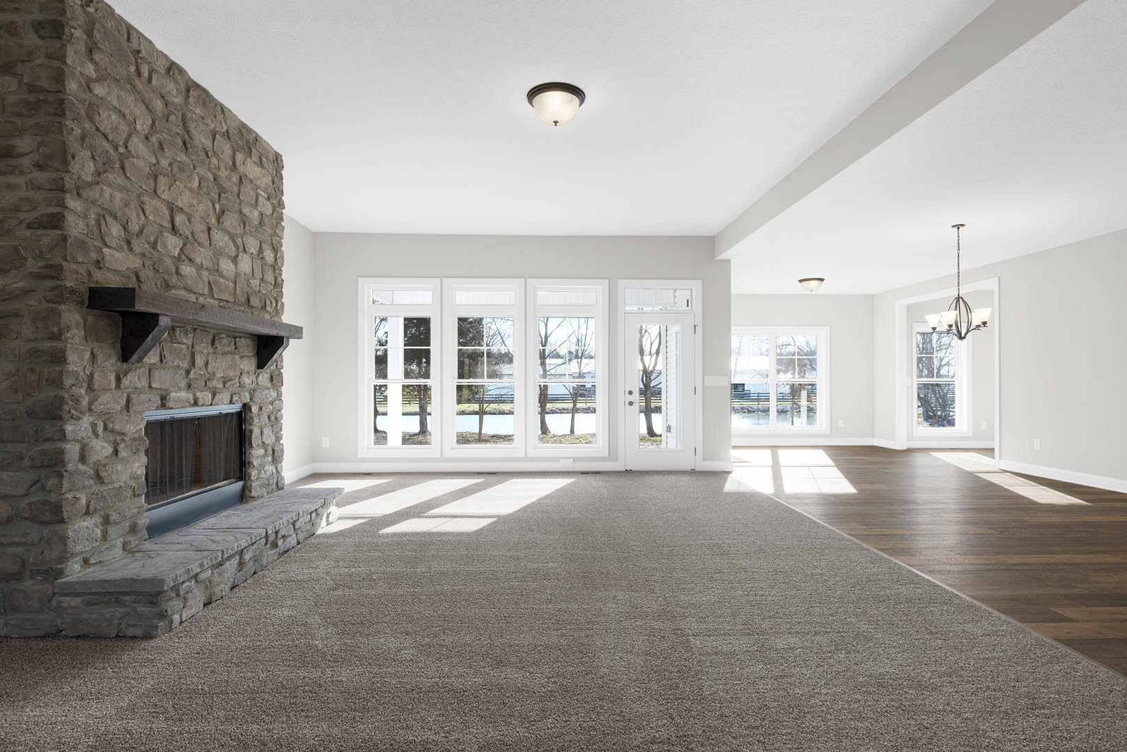 Living room with beige carpet, stone fireplace featuring a wood mantel and built-in window, large windows offering tree views, white ceiling with recessed lighting, neutral walls