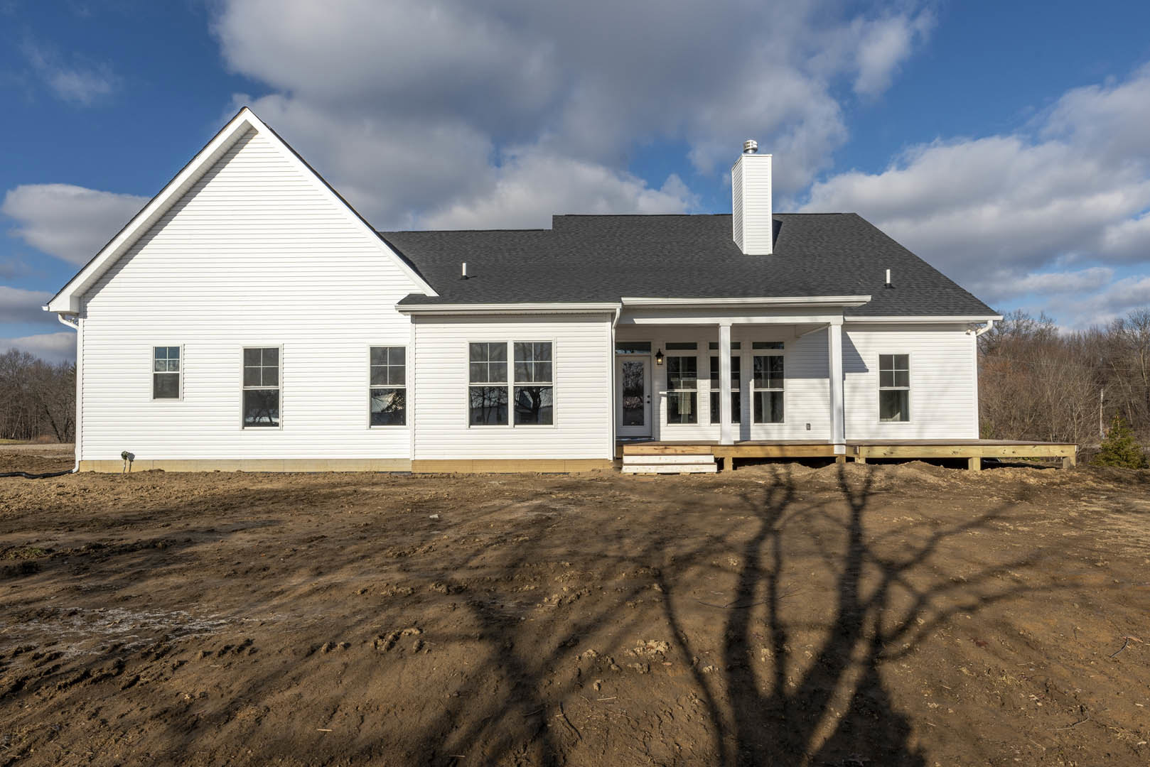 White house with black roof and white siding, large window reflecting trees, dirt yard in foreground, blue sky with scattered clouds overhead