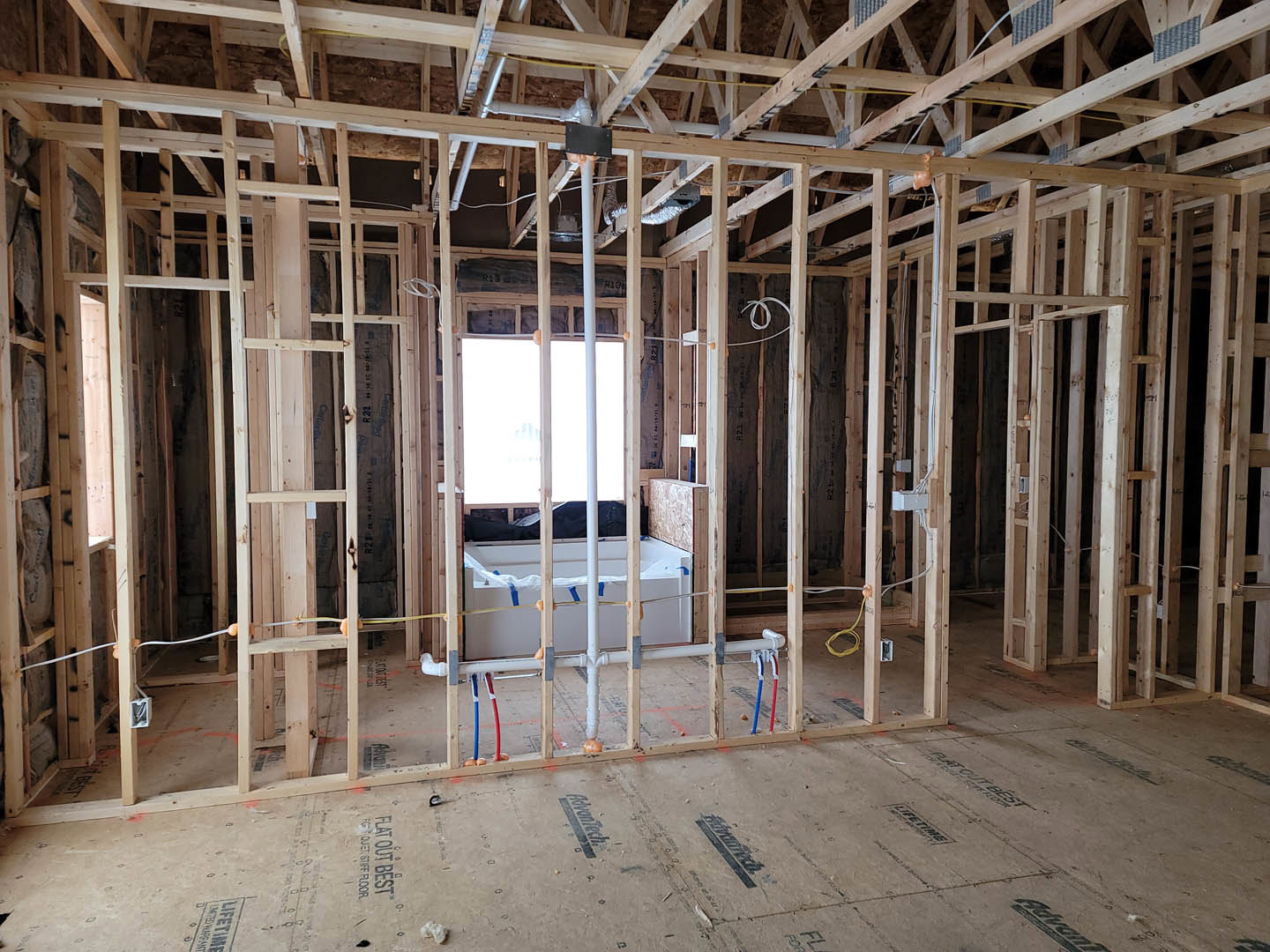 Freestanding white bathtub beneath large window, light wood flooring, white walls, exposed ceiling beams