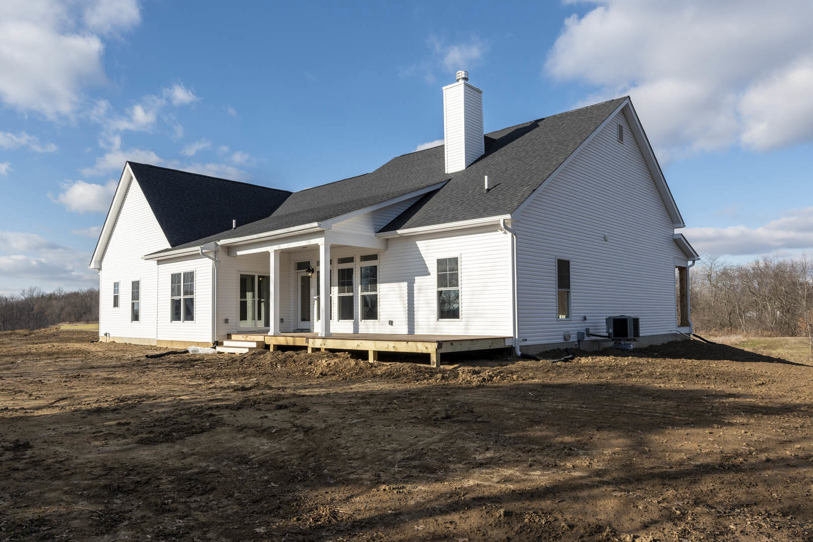 White siding house under construction with covered porch, dirt yard, and blue sky; large windows with white frames visible.