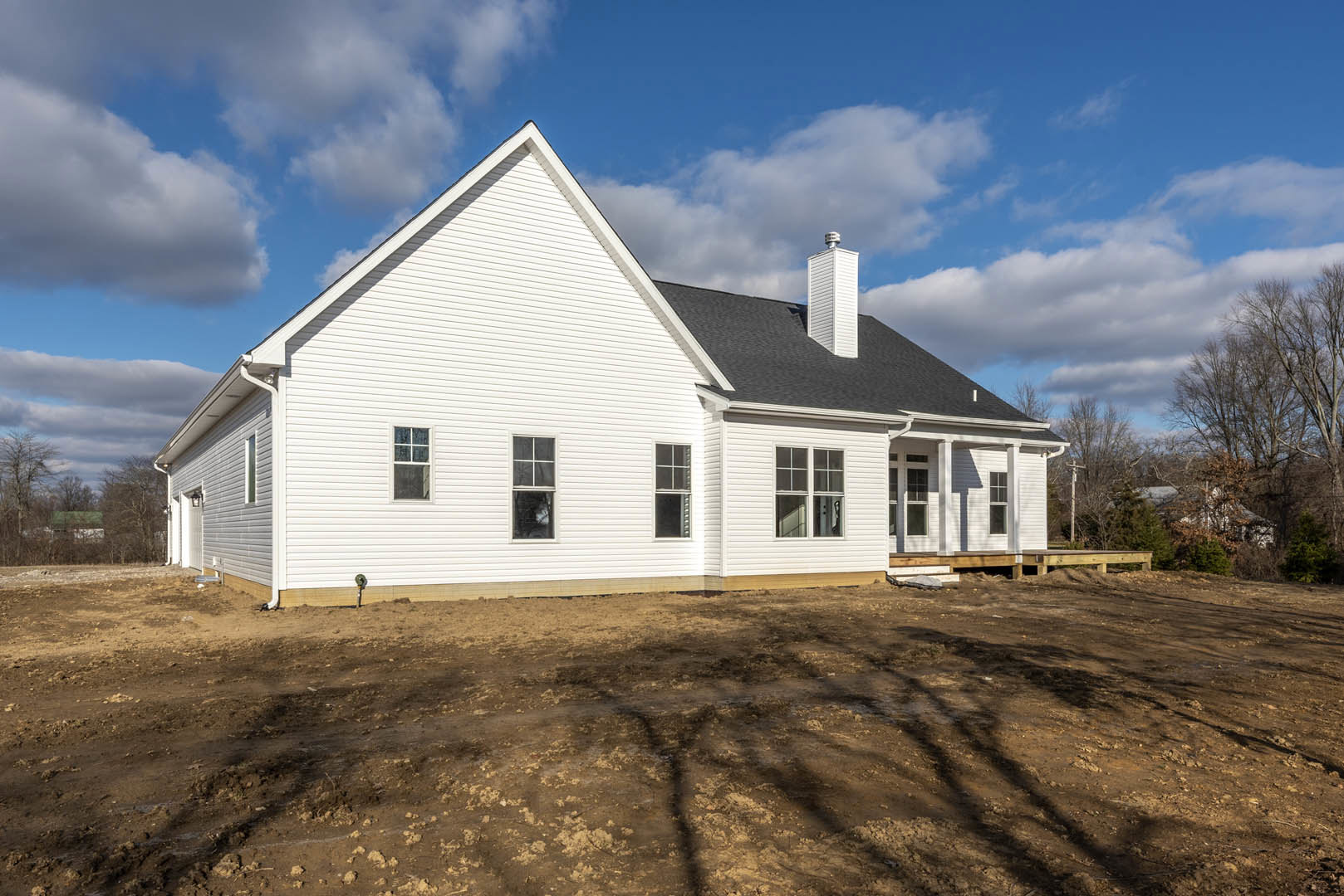 White house under construction with exposed dirt foreground, prominent chimney, white-framed windows, and scattered clouds in blue sky