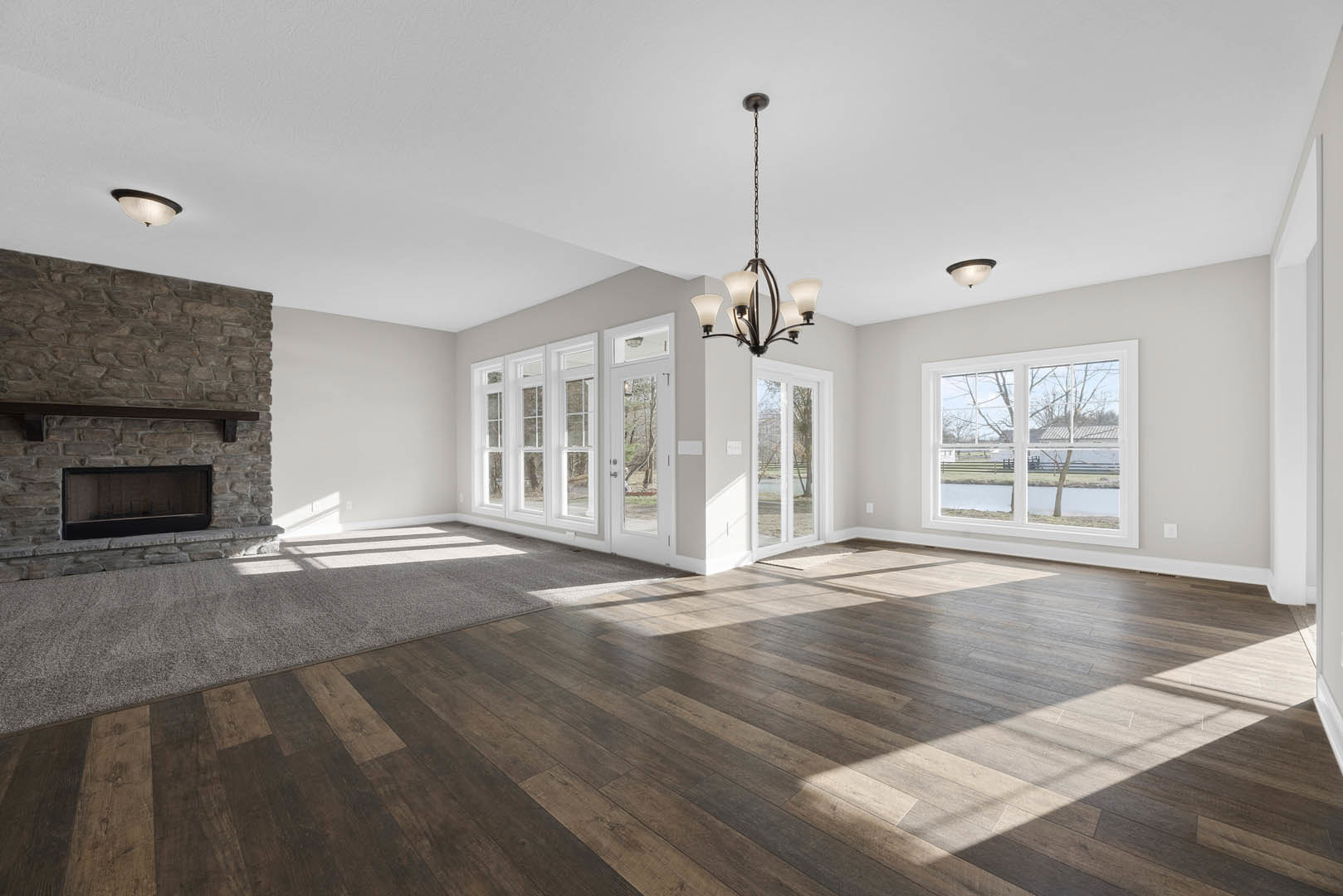 Spacious room featuring hardwood flooring, stone fireplace, large window overlooking river and farmland, chandelier, and doorway with area rug