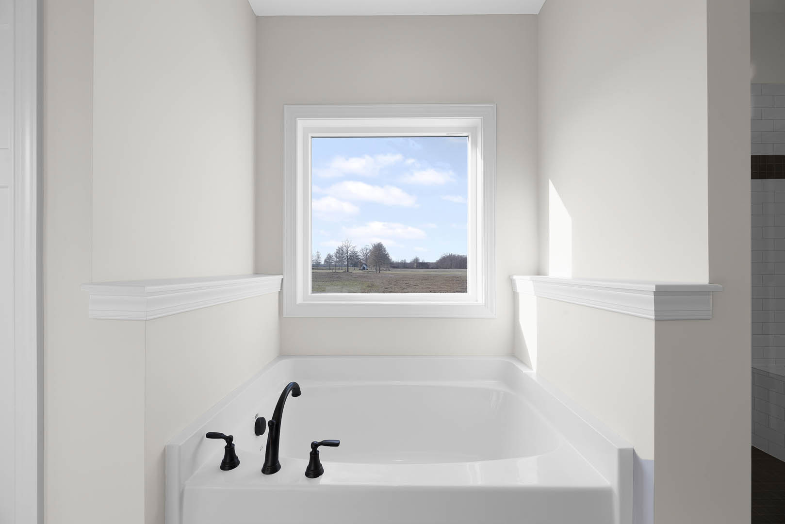 Freestanding white bathtub beneath a large window overlooking a grassy field and trees, blue sky and clouds visible, modern bathroom with neutral wall finishes