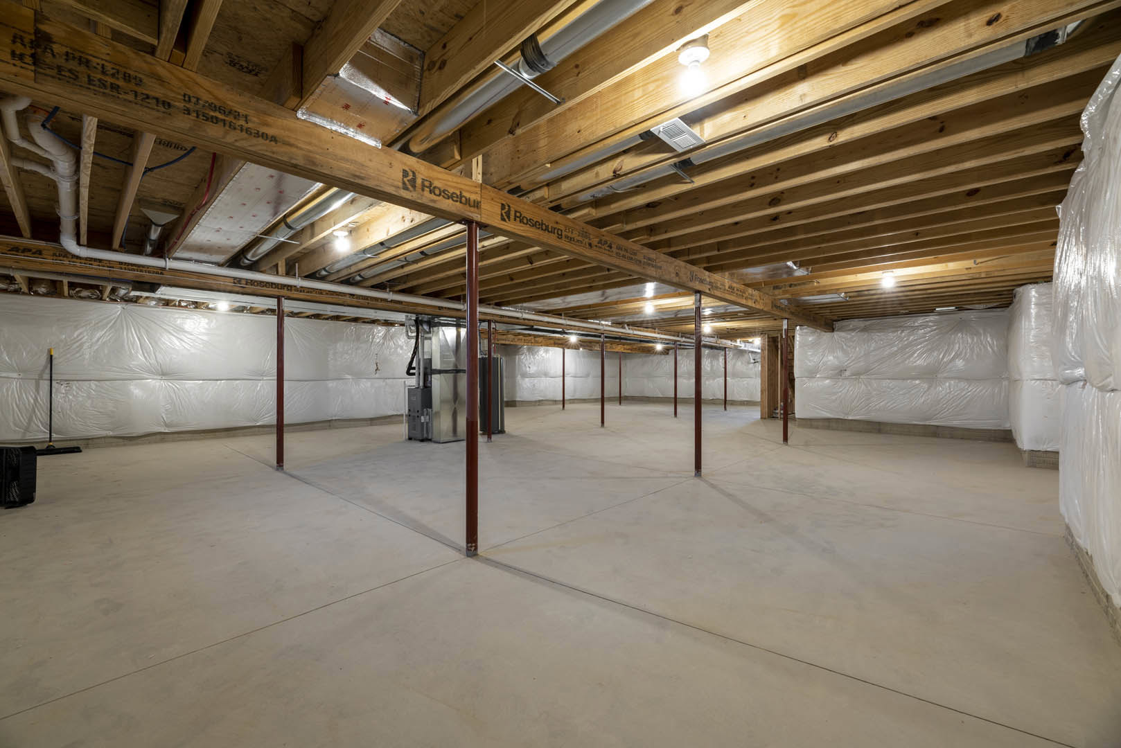 Concrete floor with red metal poles, exposed wood ceiling beams, white plastic sheeting covering one wall, light bulb hanging from ceiling, black and white object near wall.