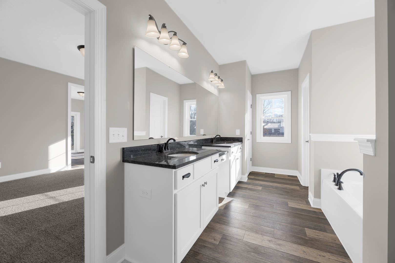 Bathroom with a wide mirror above a white sink set on black countertops, wood flooring, and a window offering a view of a neighboring house
