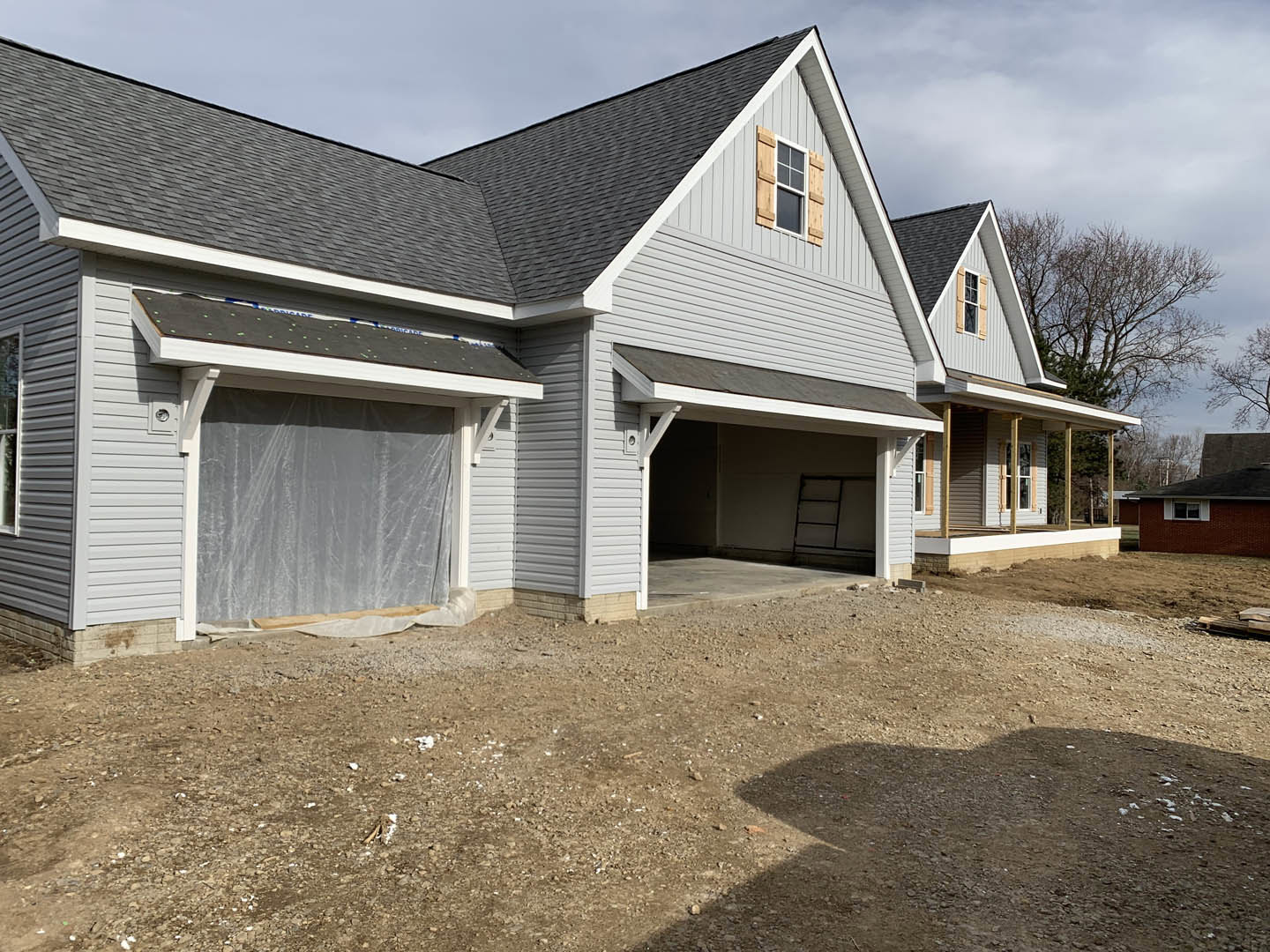 Two-story house under construction with exposed wood framing, dirt driveway, attached garage, and surrounding trees; visible window with wood shutters and white curtain, close-up