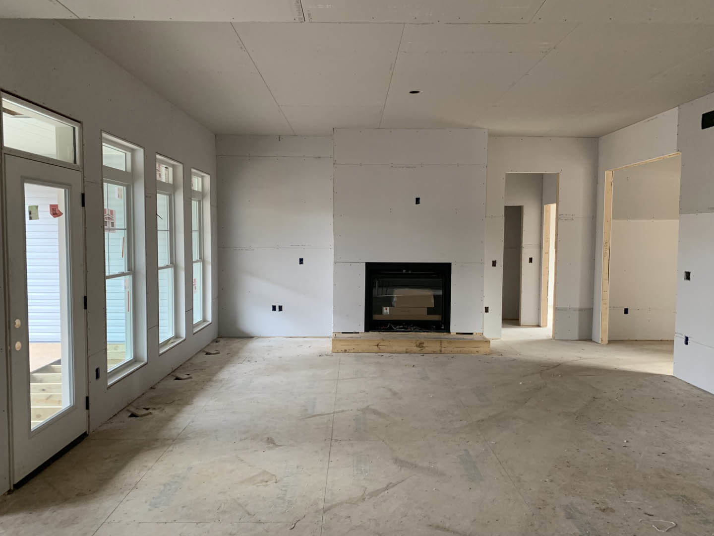 Concrete floor and white plaster walls, modern fireplace with black surround, large windows framed in wood, glass door with visible box near entry.