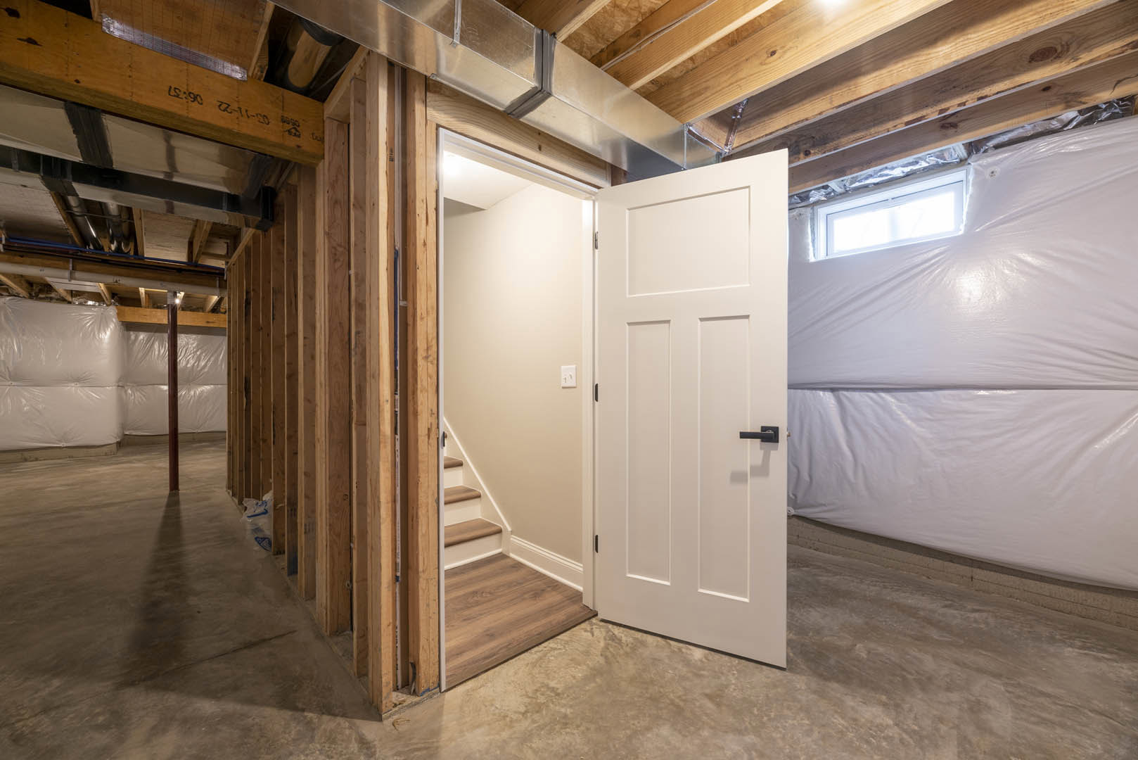 White-painted room featuring a black-handled door, white wall with light switch, staircase with wood and metal beams, and visible plastic wall covering.