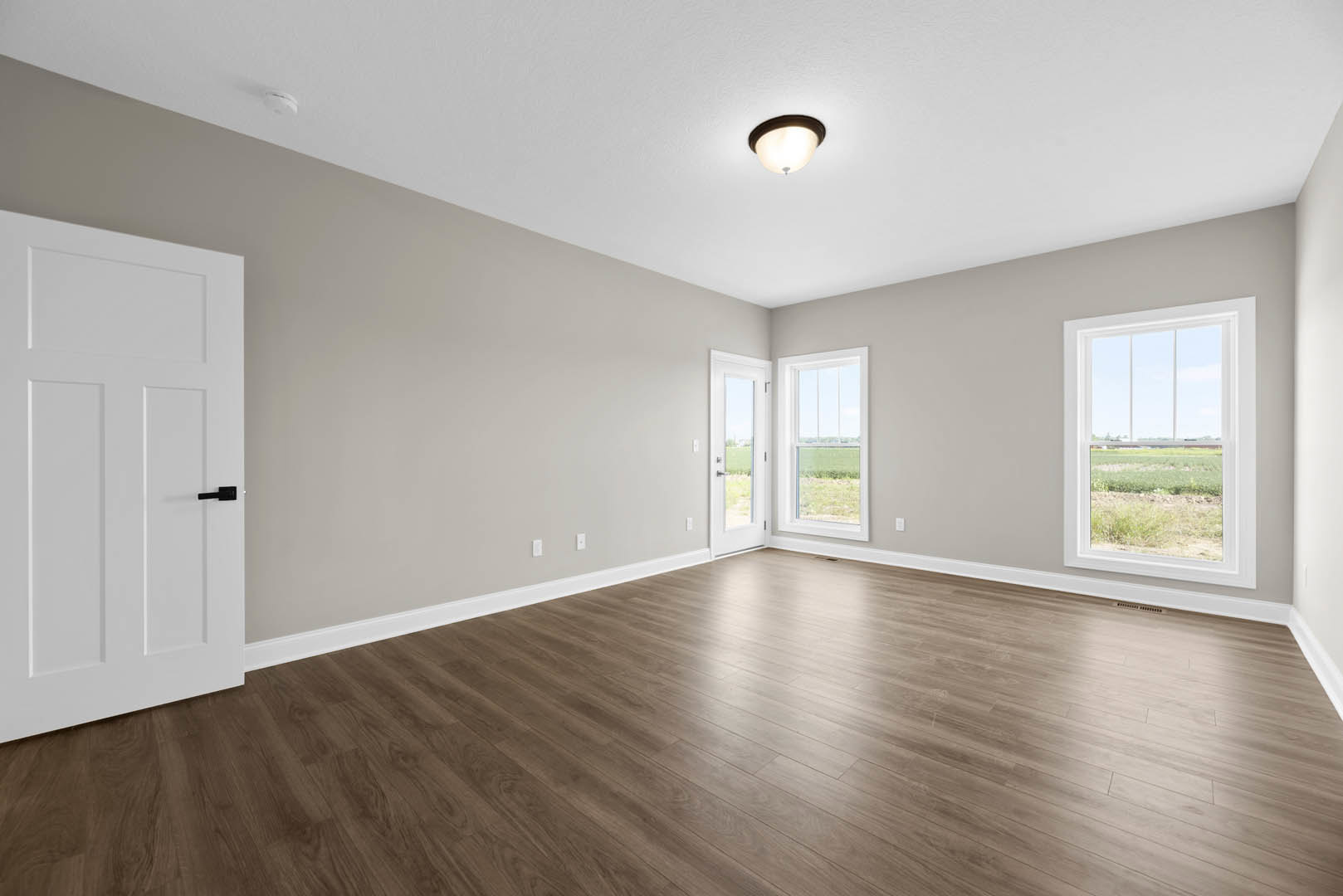 Hardwood floor room with white door featuring black handle, ceiling light fixture, and window overlooking a grassy field