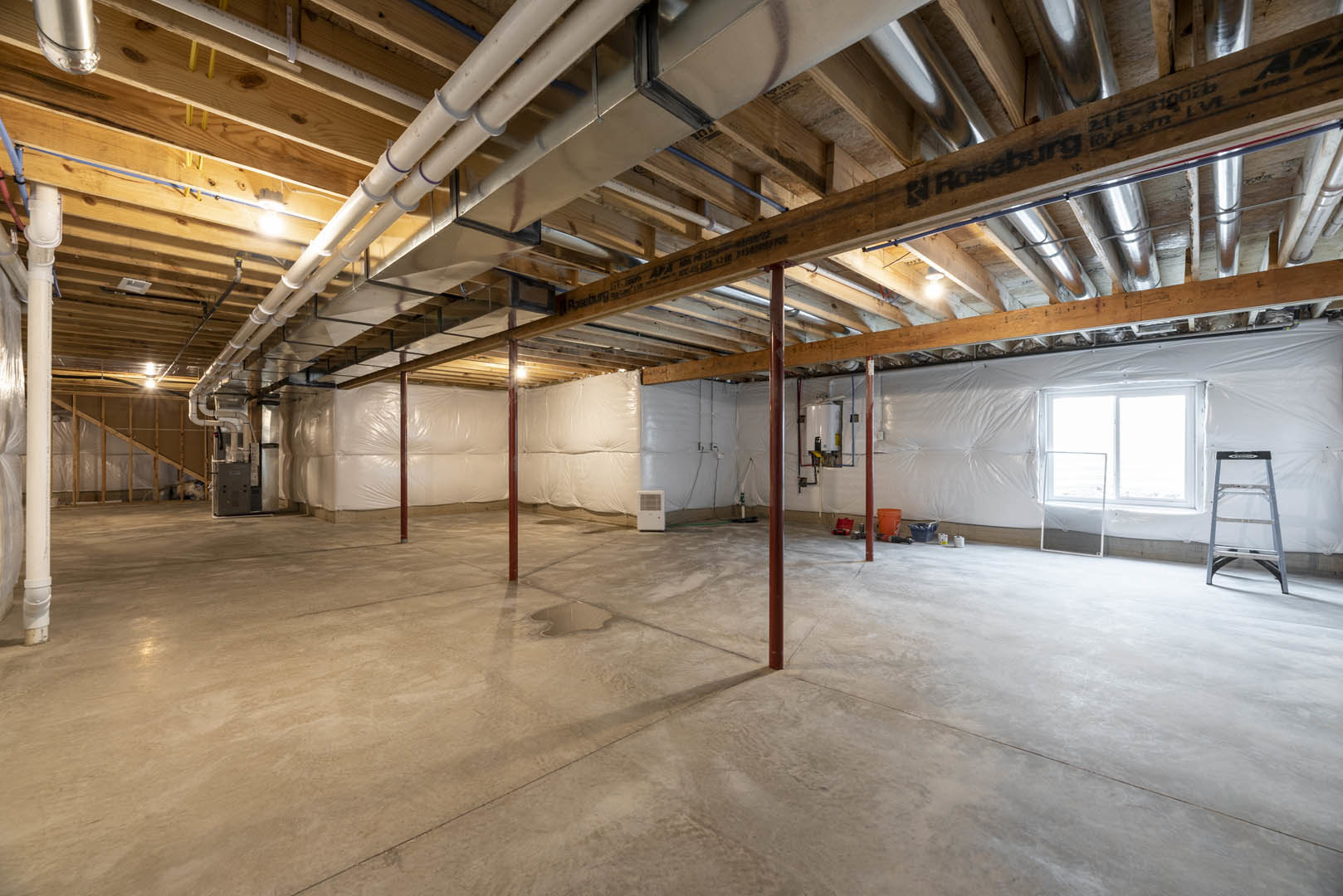 Unfinished basement with exposed ceiling pipes, white-framed window, wet concrete floor with puddles, and metal ladder against white wall