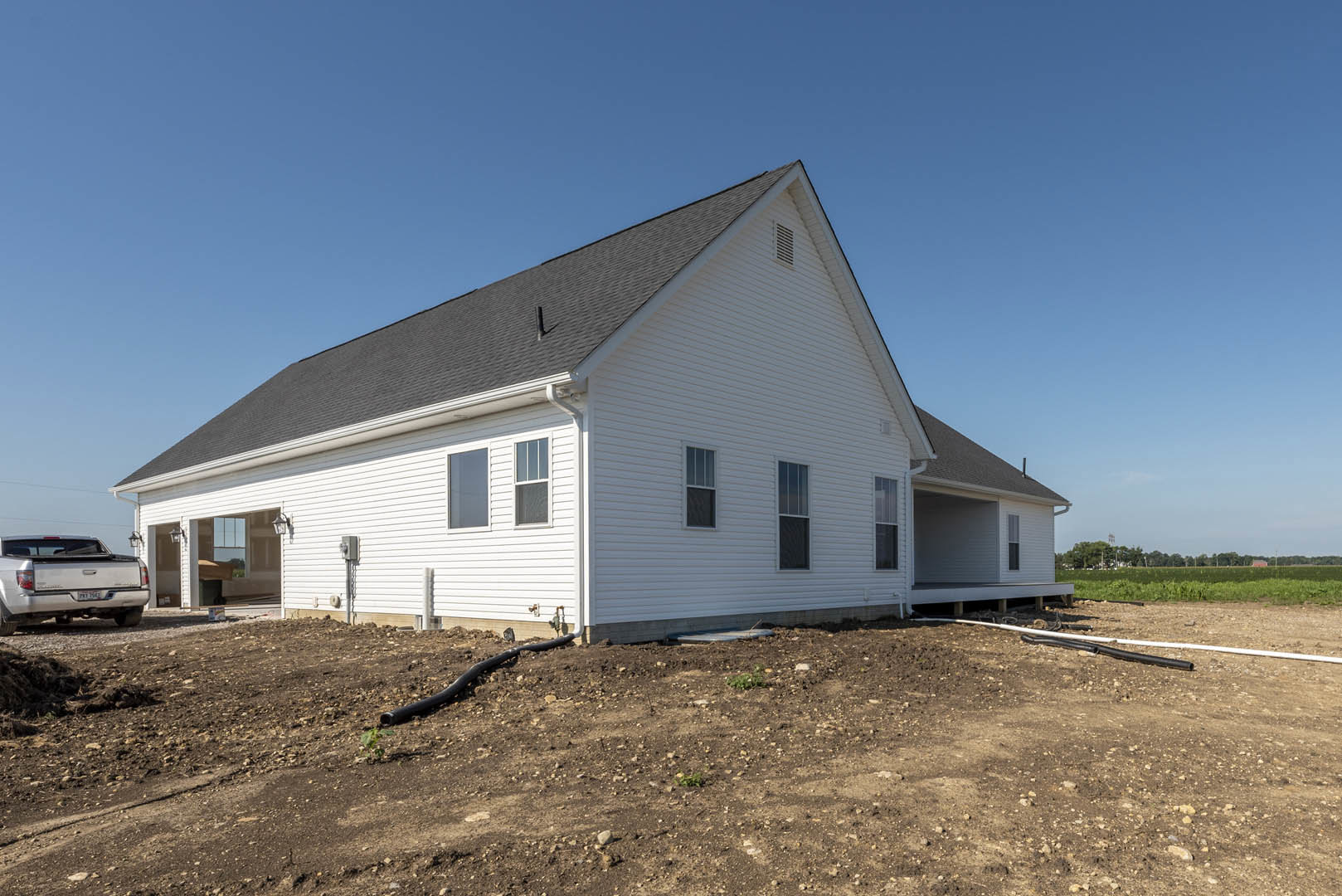 White house with black roof under construction, dirt lot with rocks and exposed pipe, silver truck parked with trunk open, blue sky overhead, window visible on building facade